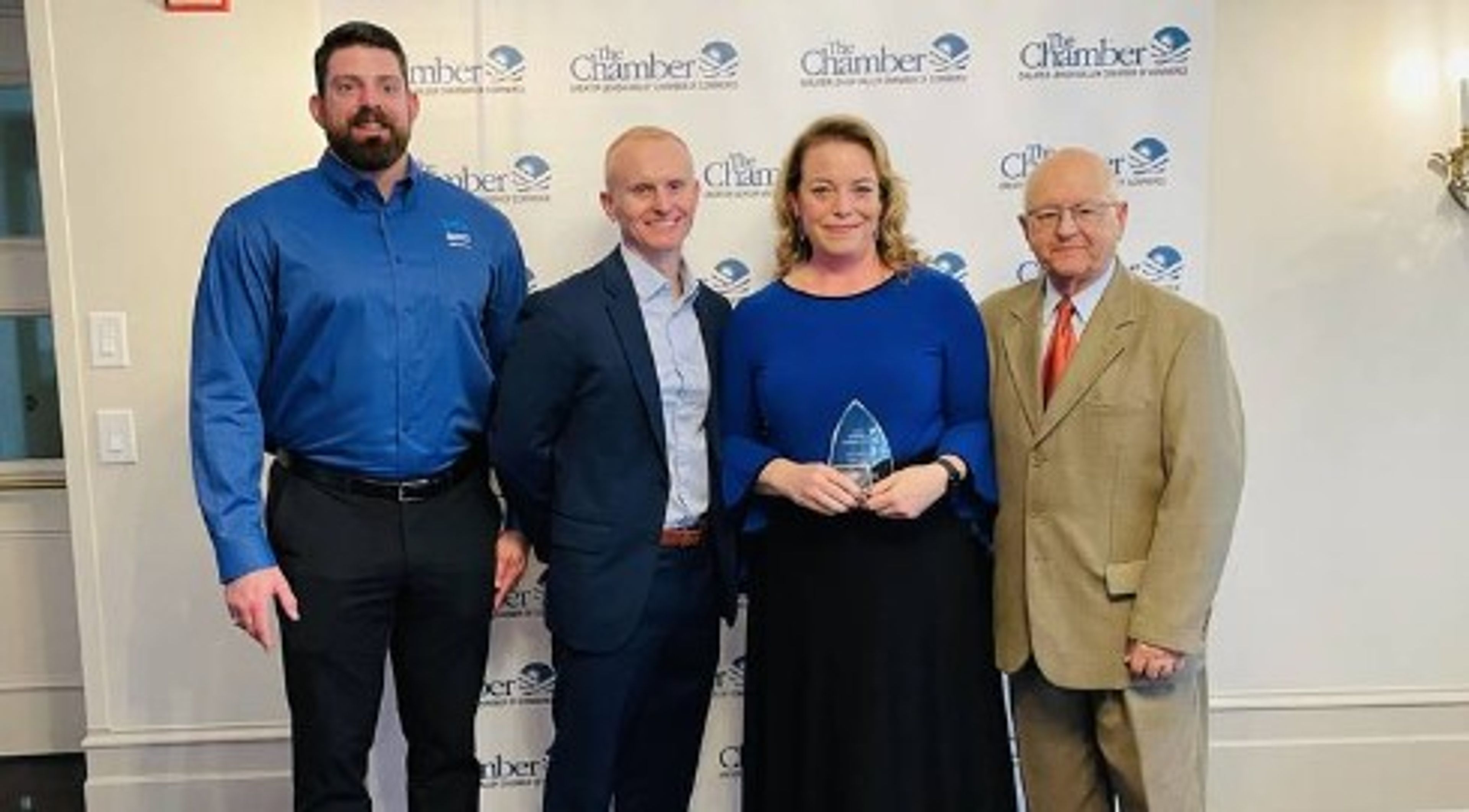 Four people stand together at an awards event. The woman in the center holds a glass trophy. A banner behind them reads “The Chamber.” All are dressed in business attire and smiling at the camera.