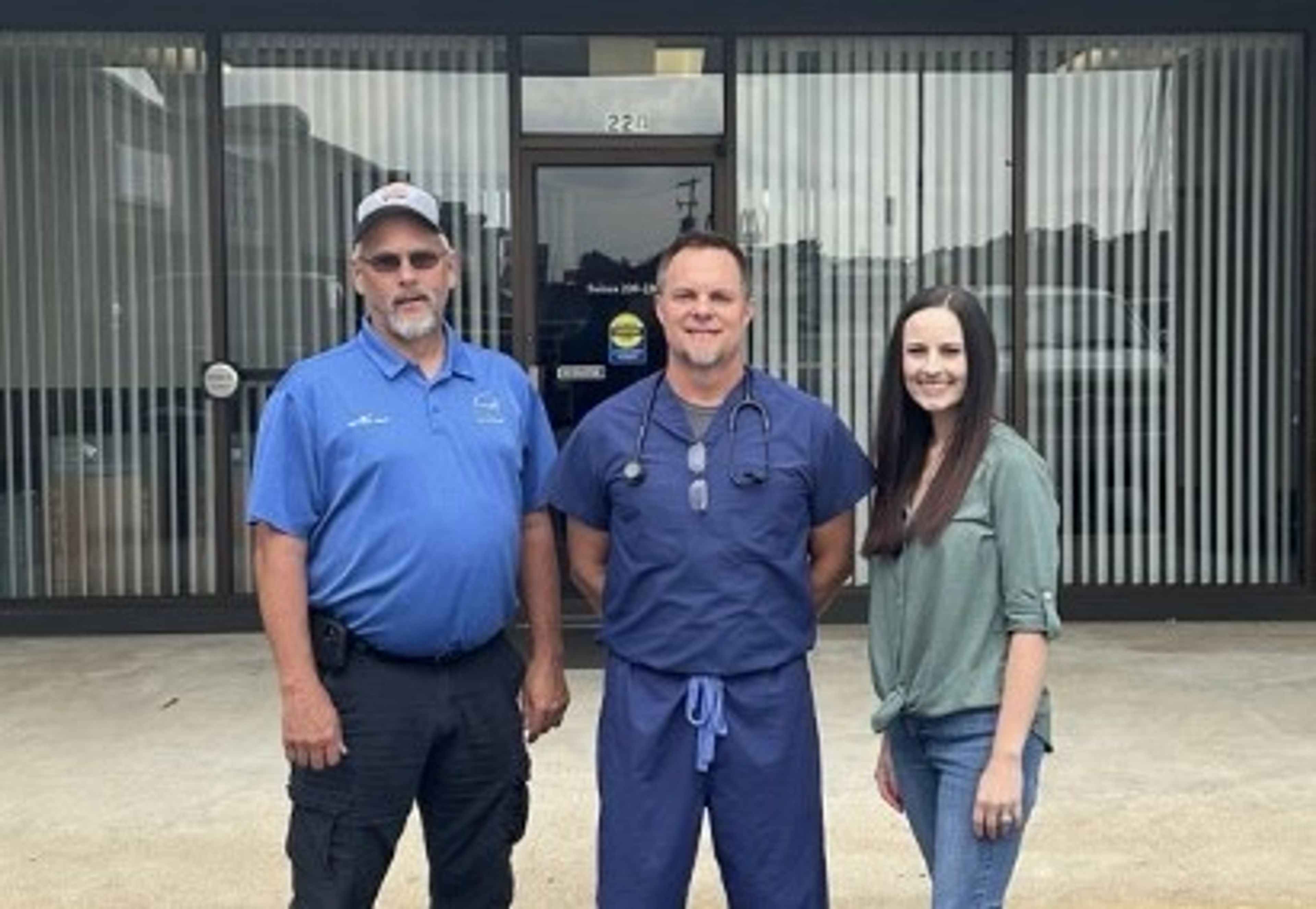 Three people stand smiling in front of a building entrance. One wears a blue polo and hat, one is in medical scrubs with a stethoscope, and one wears a green blouse and jeans. Vertical blinds cover the windows behind them.