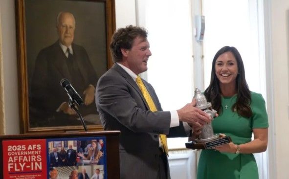 A man in a suit presents a silver fire hydrant trophy to a smiling woman in a green dress at an event, with a portrait and a “2025 AFS Government Affairs Fly-In” sign visible.