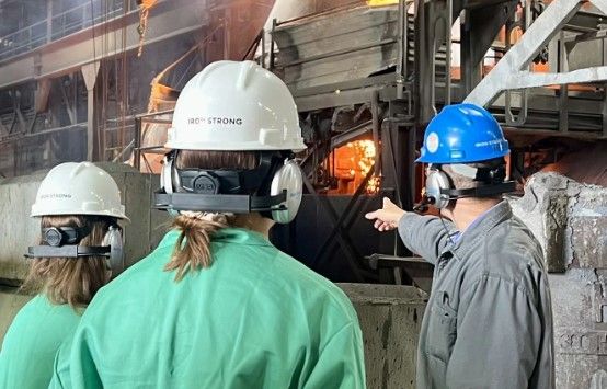 Three people in protective gear and helmets observe a glowing industrial furnace. One person points toward the furnace, which emits bright orange light, while the others watch attentively.