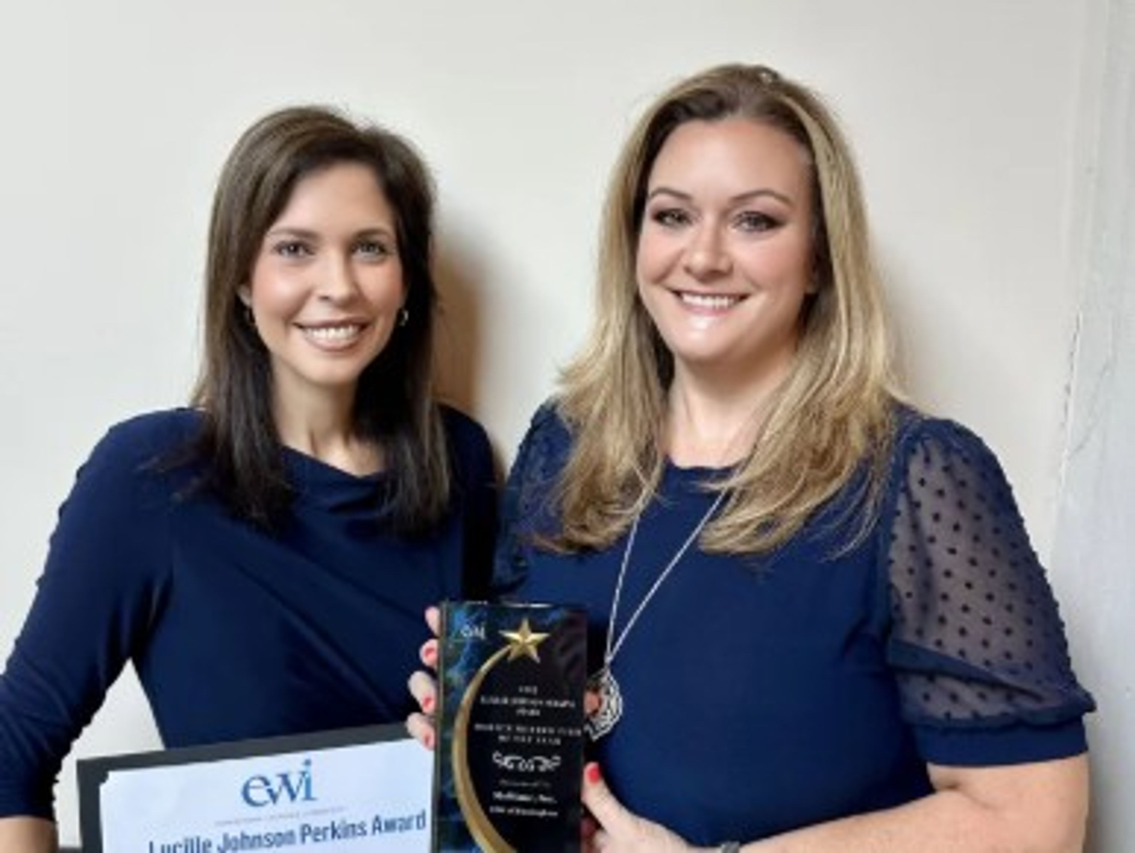 Two women smile while holding an award and a certificate. Both wear navy tops, and stand against a plain light-colored wall. The woman on the left holds a framed certificate; the woman on the right holds a star-shaped trophy.