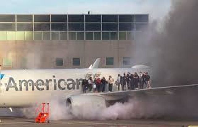 A group of people stand on the wing of an airplane surrounded by smoke, appearing to evacuate. The plane is near a building and the word “American” is visible on the aircraft.