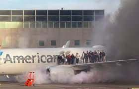 A group of people stand on the wing of an airplane surrounded by smoke, appearing to evacuate. The plane is near a building and the word “American” is visible on the aircraft.