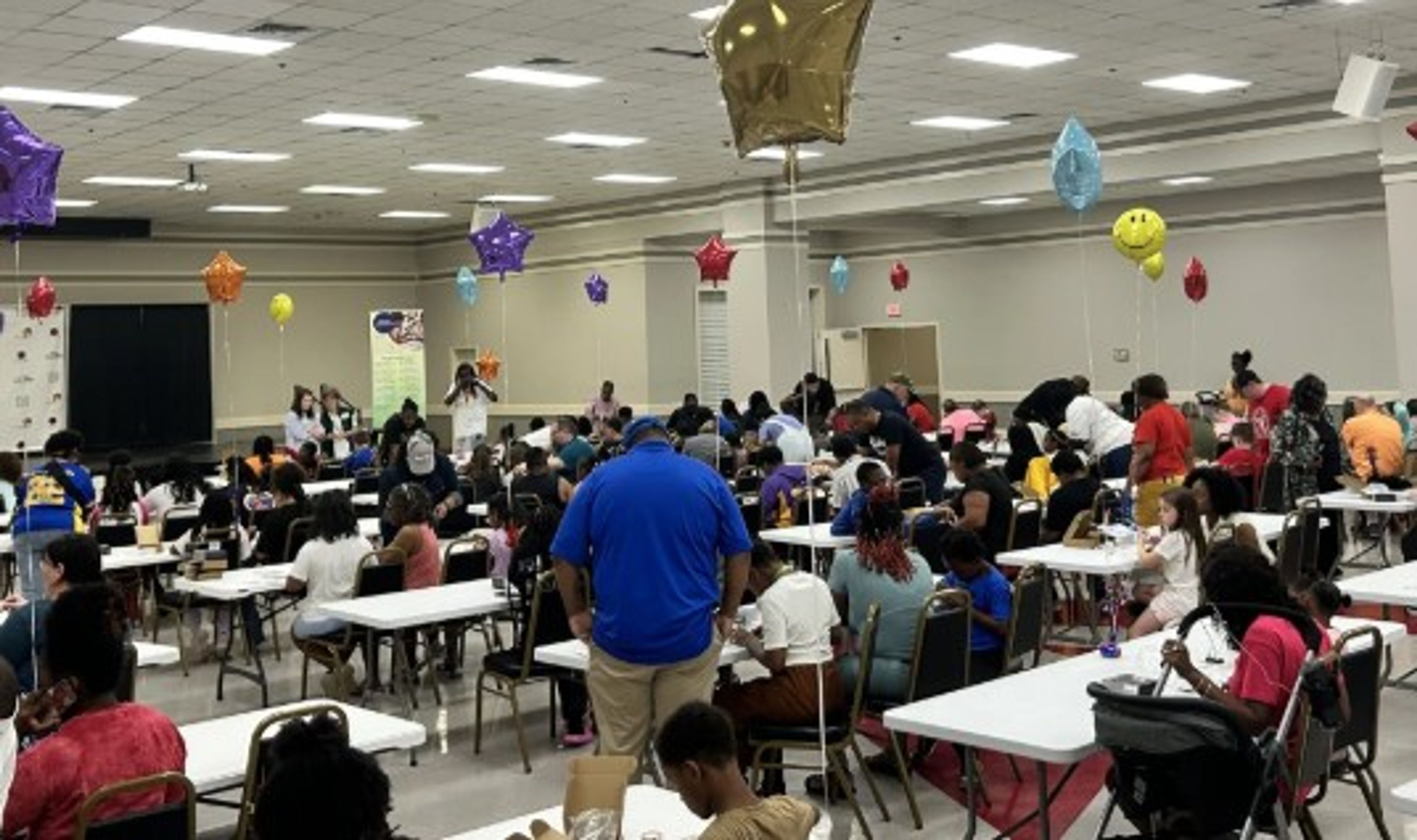 A large group of people, including many children, sit at tables in a decorated hall with colorful balloons, attending an event or gathering.