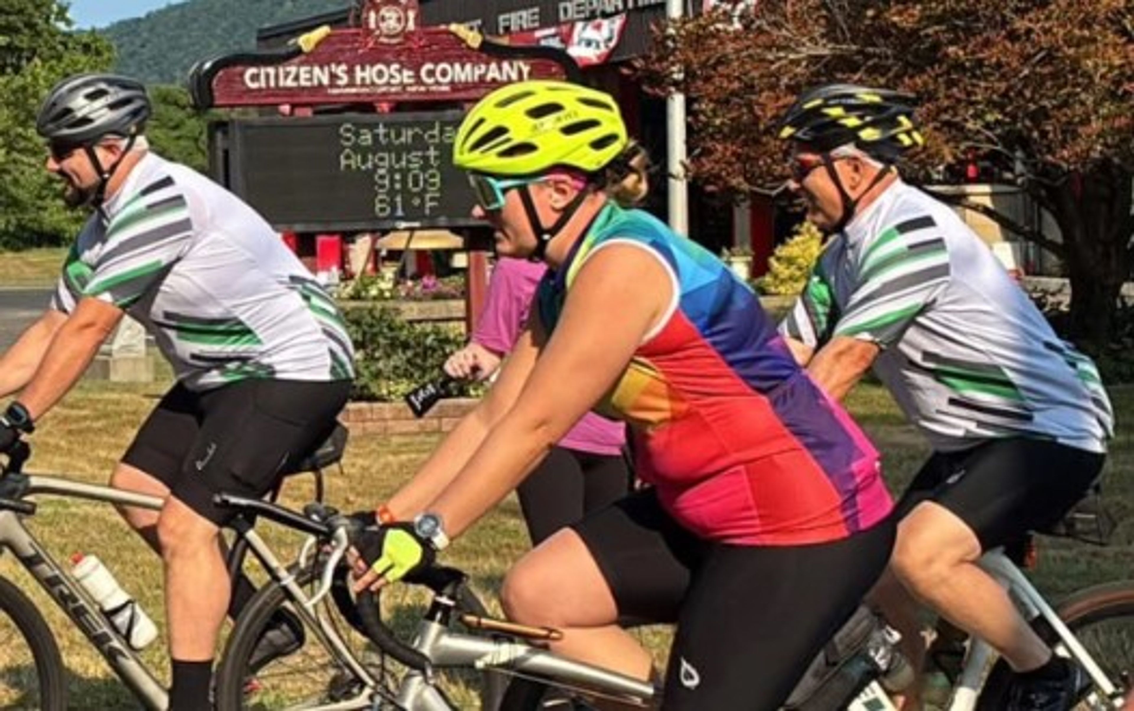 Three cyclists ride past a fire station sign on a sunny day. Two wear matching white and green jerseys and helmets; one wears a colorful jersey and yellow helmet. Trees and a digital sign are visible in the background.