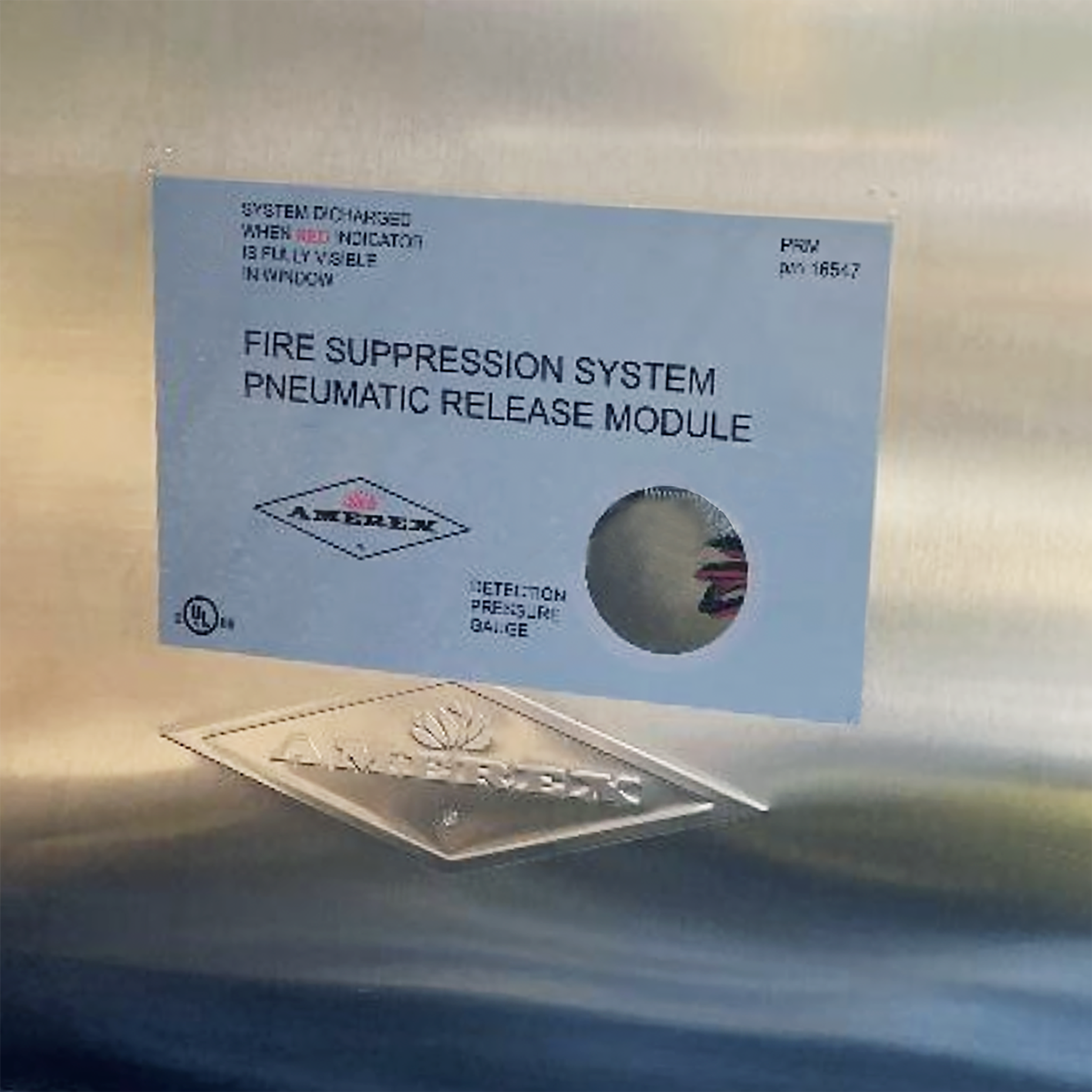 A label on a metallic surface reads “FIRE SUPPRESSION SYSTEM PNEUMATIC RELEASE MODULE” with instructions and official logos. Part of a circular gauge is visible through a cutout.