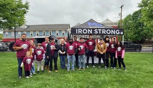 A group of people wearing matching maroon shirts pose together on grass in front of a black “IRON STRONG” tent, with buildings and trees in the background on a cloudy day.
