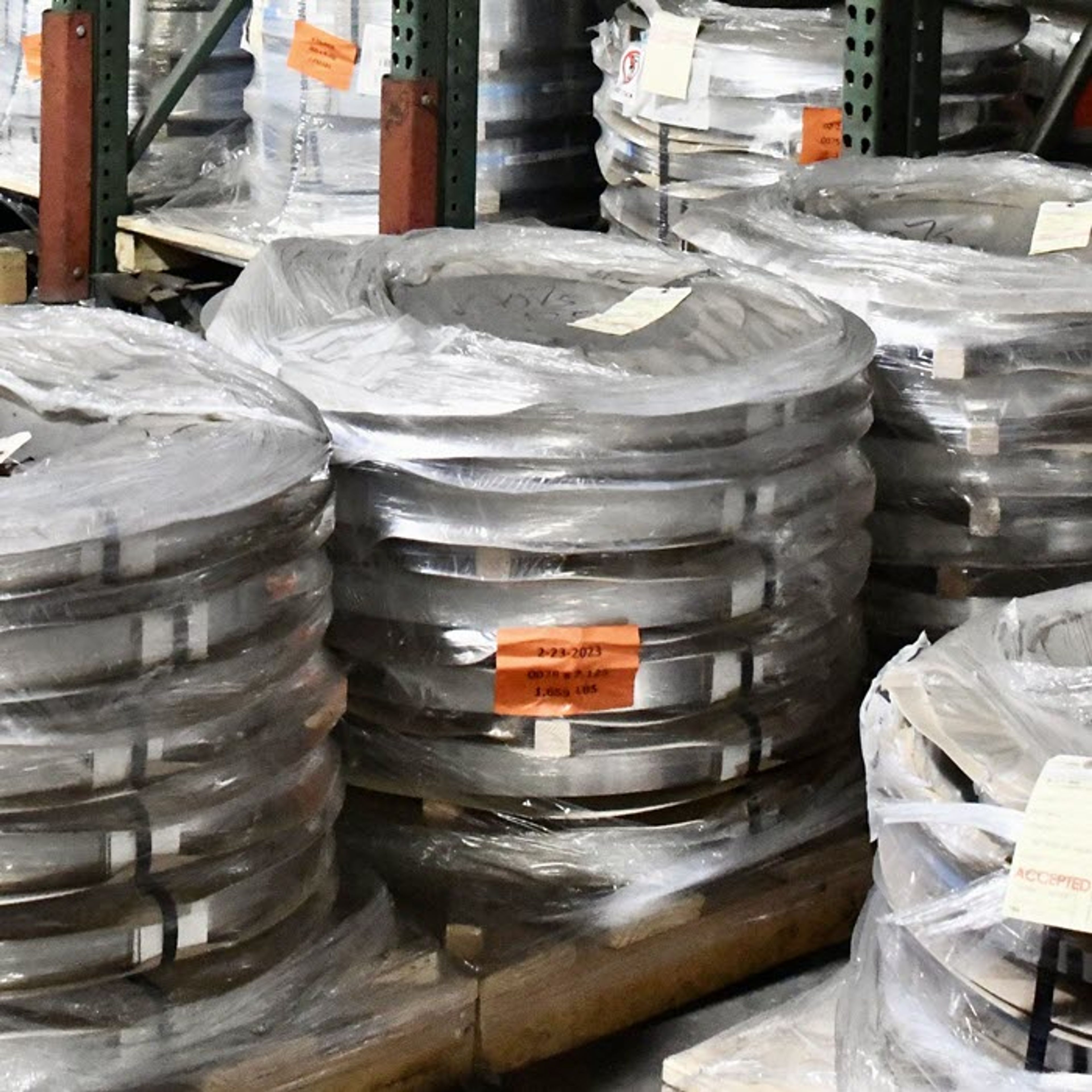 Stacks of coiled metal strips wrapped in plastic sit on wooden pallets in an industrial warehouse, surrounded by shelves and various materials.