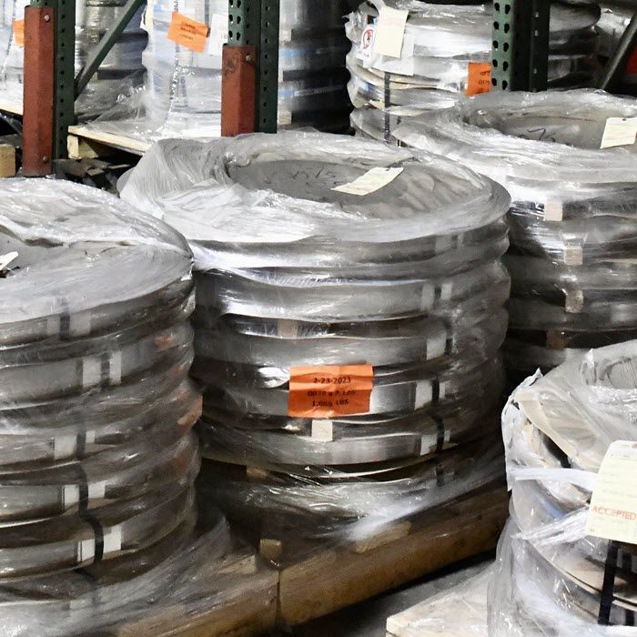 Stacks of coiled metal strips wrapped in plastic sit on wooden pallets in an industrial warehouse, surrounded by shelves and various materials.