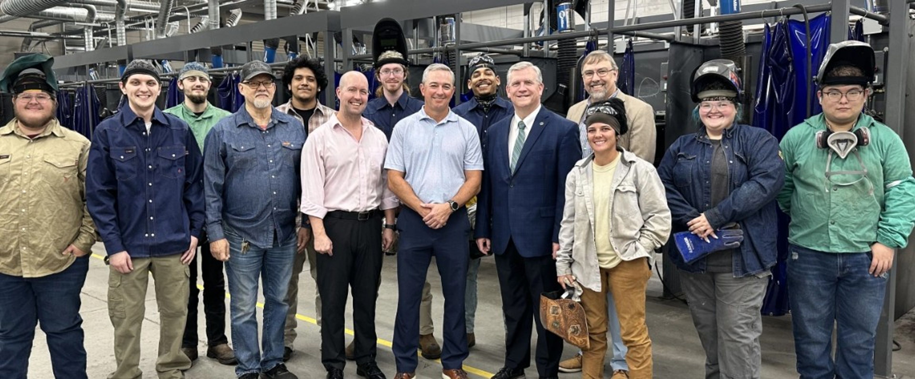 A group of people, including students in work attire and adults in business clothes, pose and smile together inside an industrial workshop with welding booths in the background.
