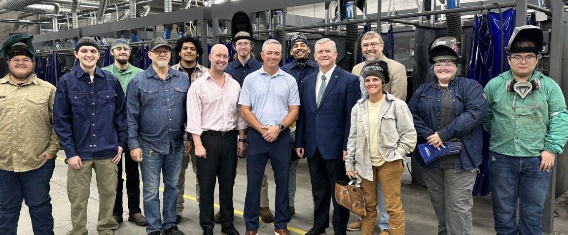A group of people, including students in work attire and adults in business clothes, pose and smile together inside an industrial workshop with welding booths in the background.
