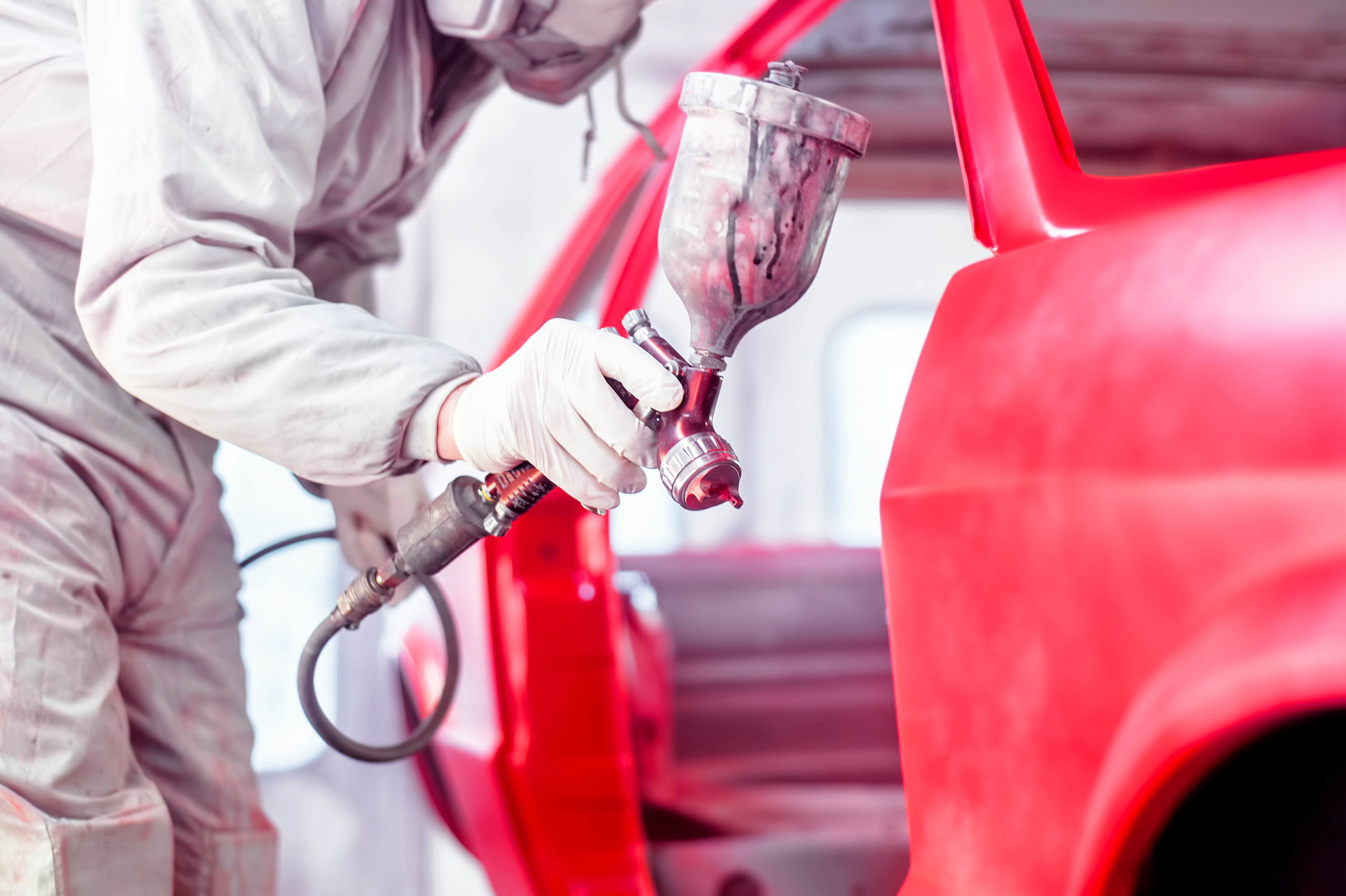 Person in protective gear spray paints a car red using a paint sprayer, focusing on the vehicle’s side in an auto body shop.