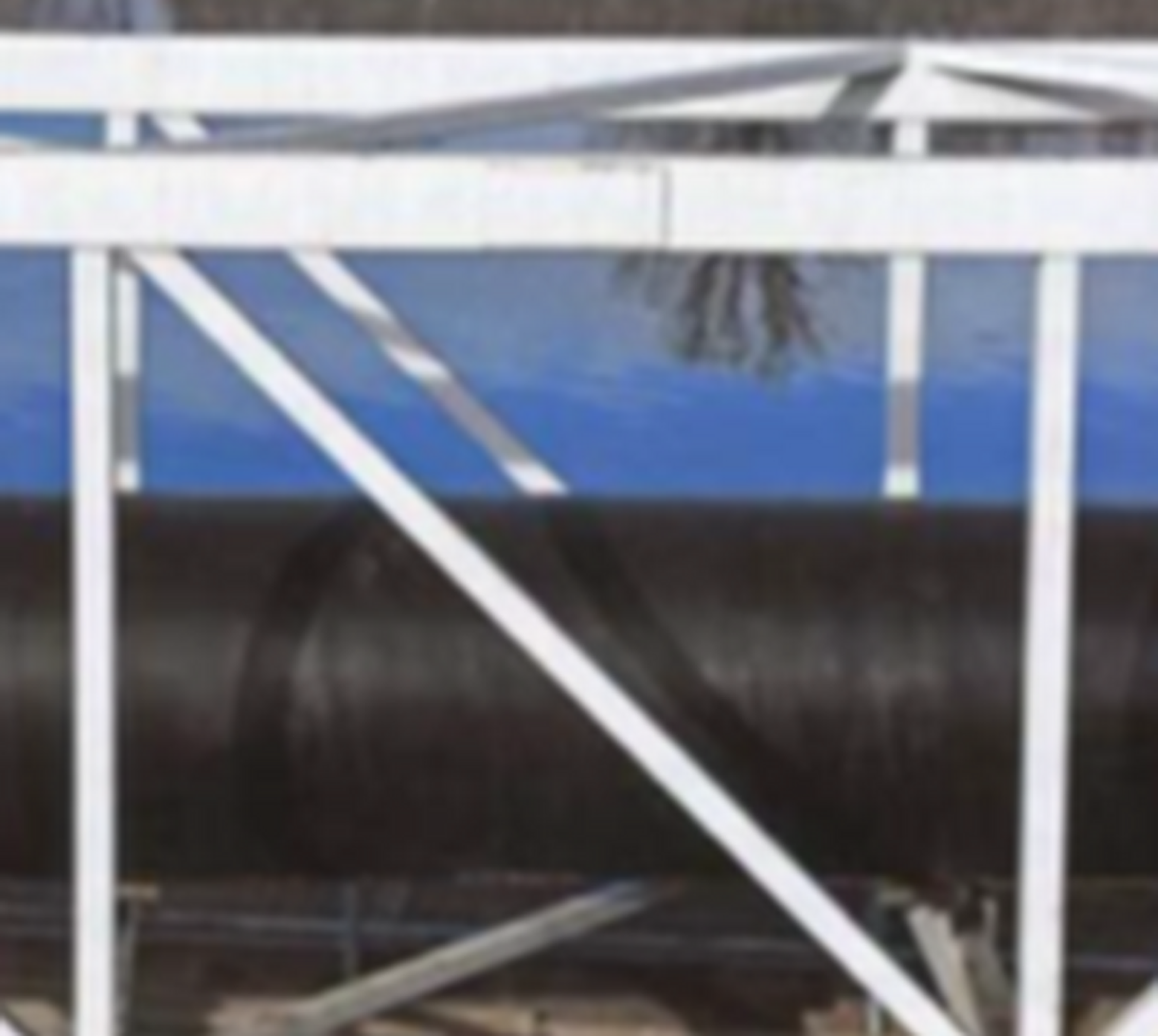 A large black pipe runs horizontally behind white metal framework with diagonal supports; blue sky and trees are reflected in a background surface, possibly water or glass.