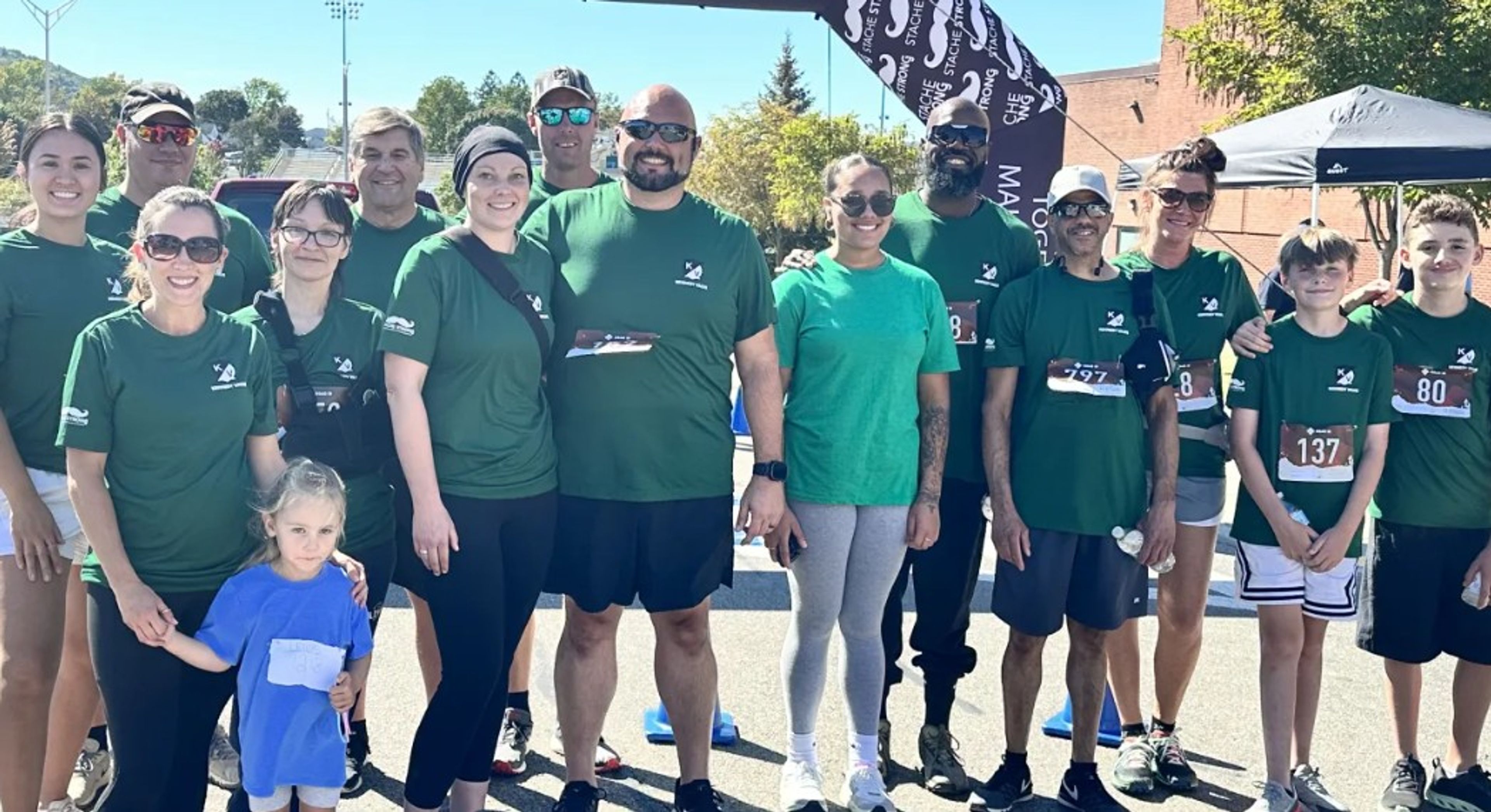 A group of people in green shirts pose together outdoors at a race event, some wearing bib numbers. A young girl in a blue shirt stands in front. Trees, tents, and a finish line banner are visible in the background.