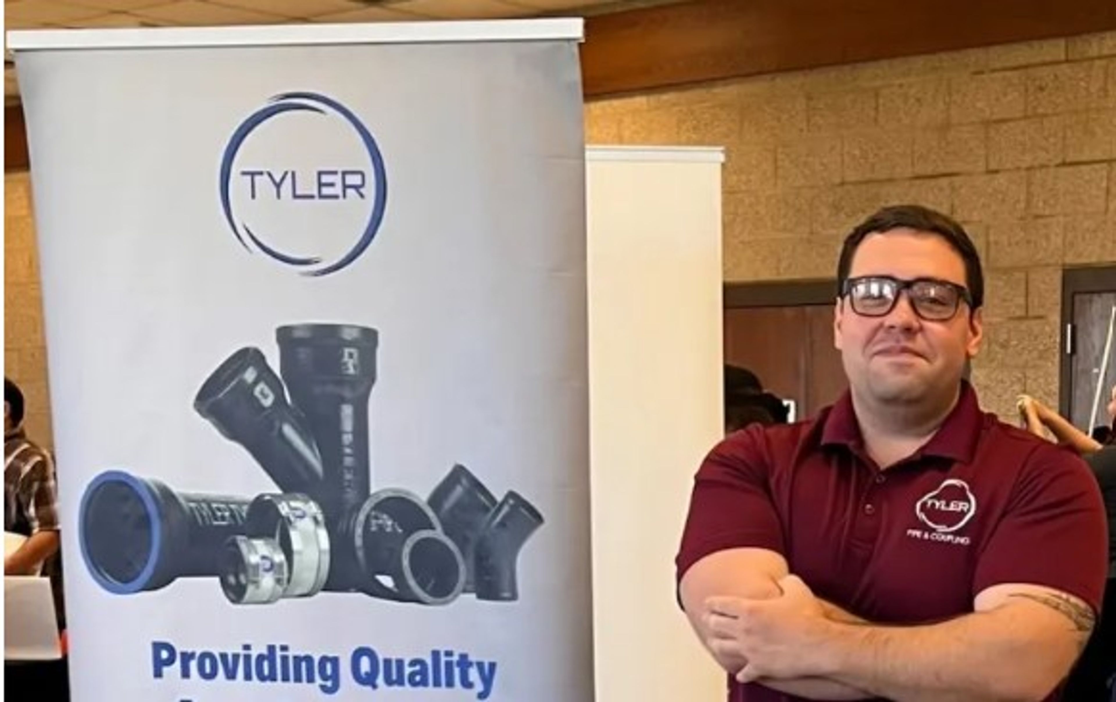 A man in glasses and a maroon shirt stands next to a “Tyler” banner displaying plumbing pipes and the phrase “Providing Quality.” The setting appears to be an indoor event or expo.