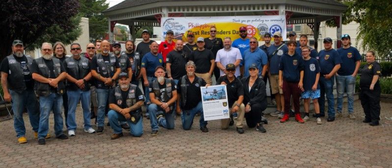 A group of men and boys pose together outdoors in front of a gazebo and a banner for First Responders Day, some wearing matching vests and uniforms, with one holding a poster.