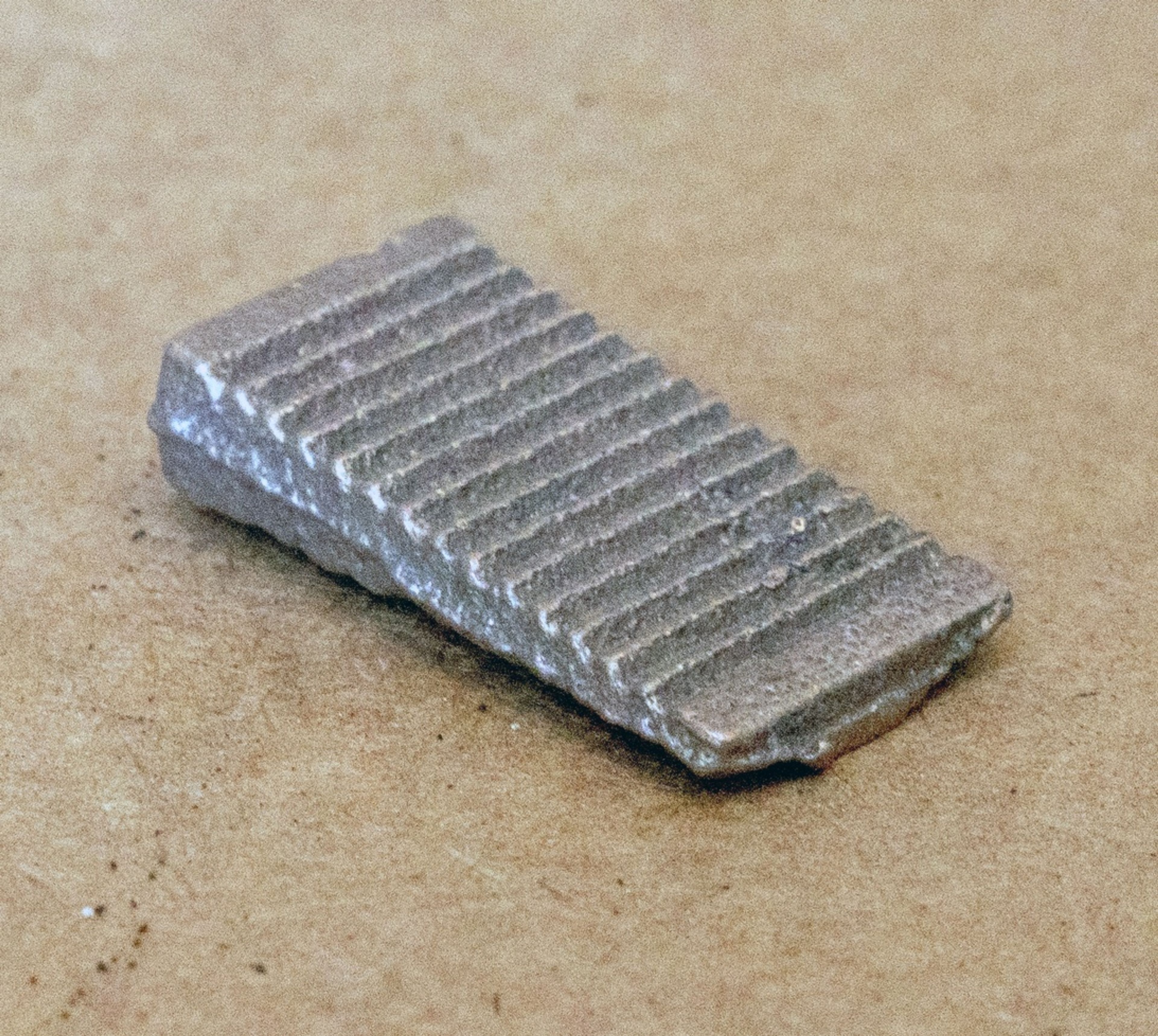 A small, wedge-shaped metal object with ridges on top, resting on a brown textured surface.