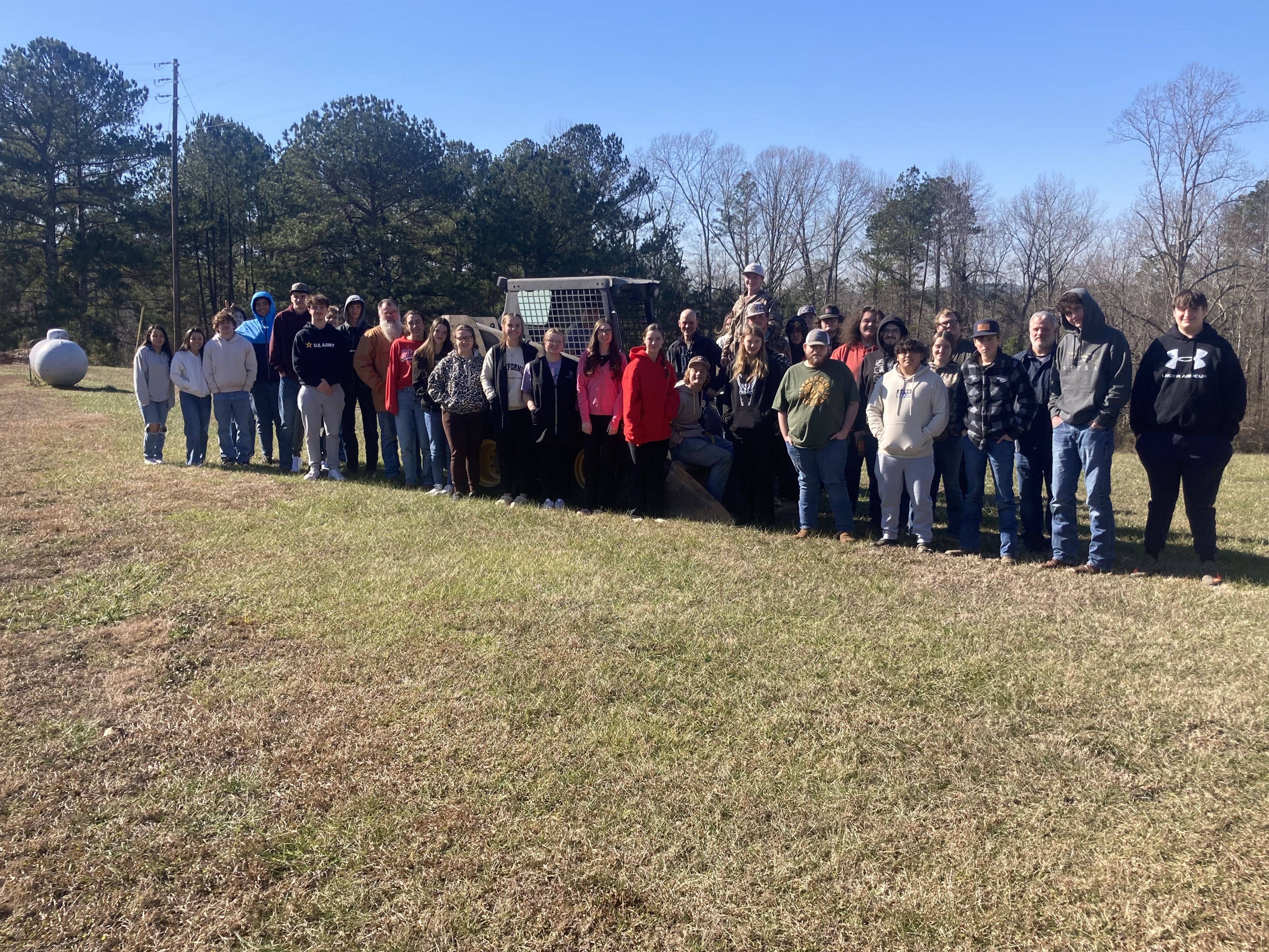 A large group of people stand together outdoors on grass, with trees and a wire structure in the background, posing for a group photo on a clear, sunny day.