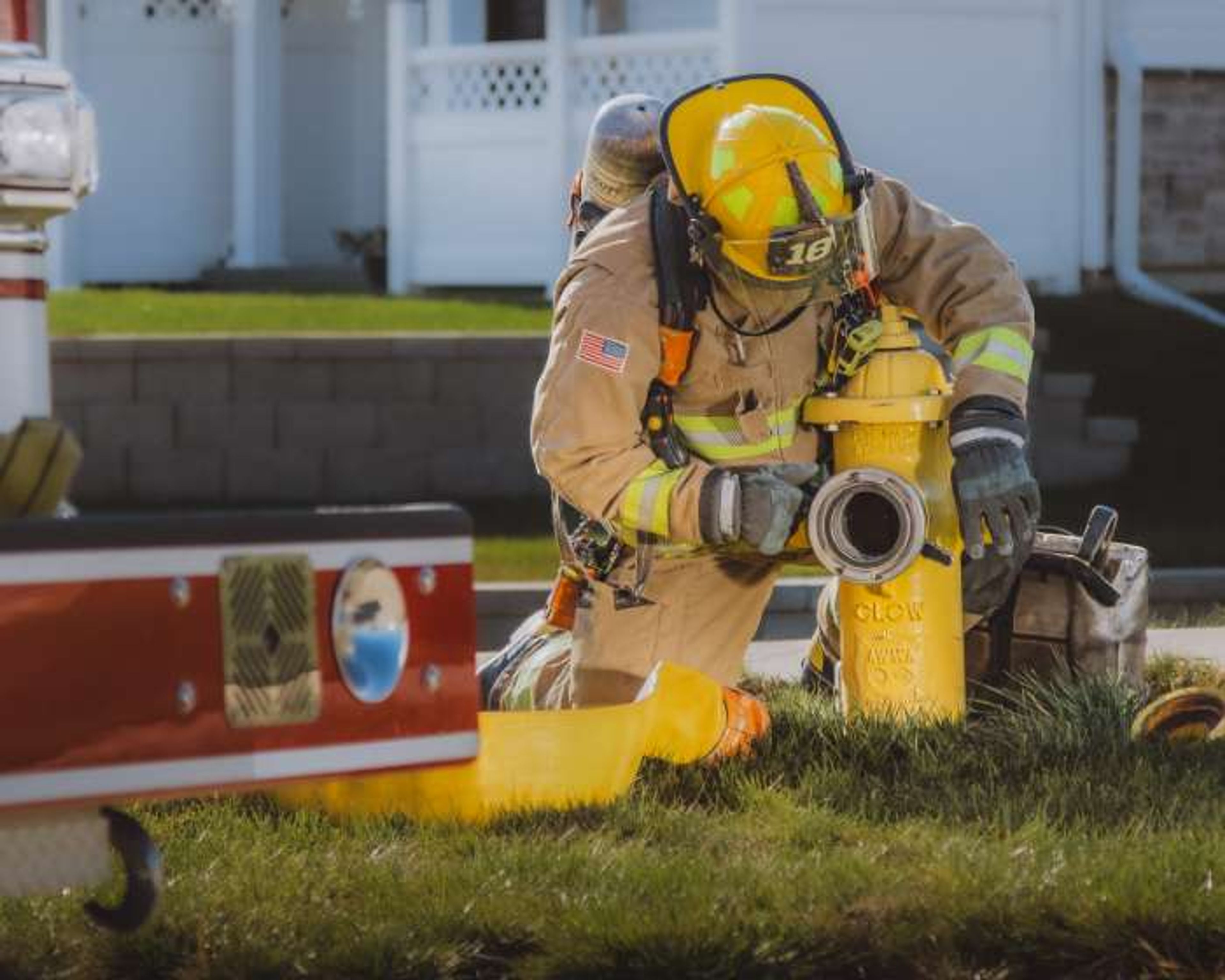 A firefighter in full gear attaches a hose to a yellow fire hydrant on a grassy area, with a fire truck and houses visible in the background.