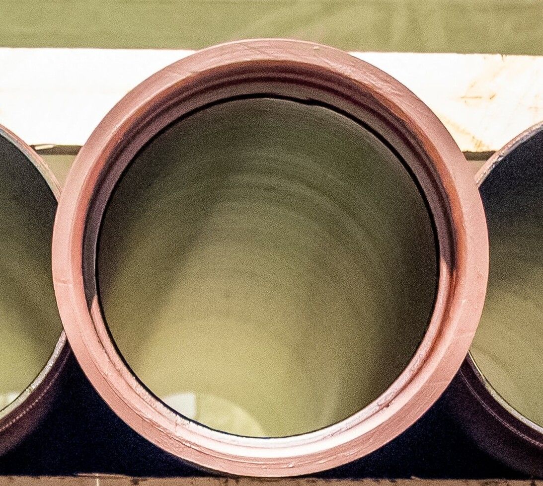 A large black pipe runs horizontally behind white metal framework with diagonal supports; blue sky and trees are reflected in a background surface, possibly water or glass.