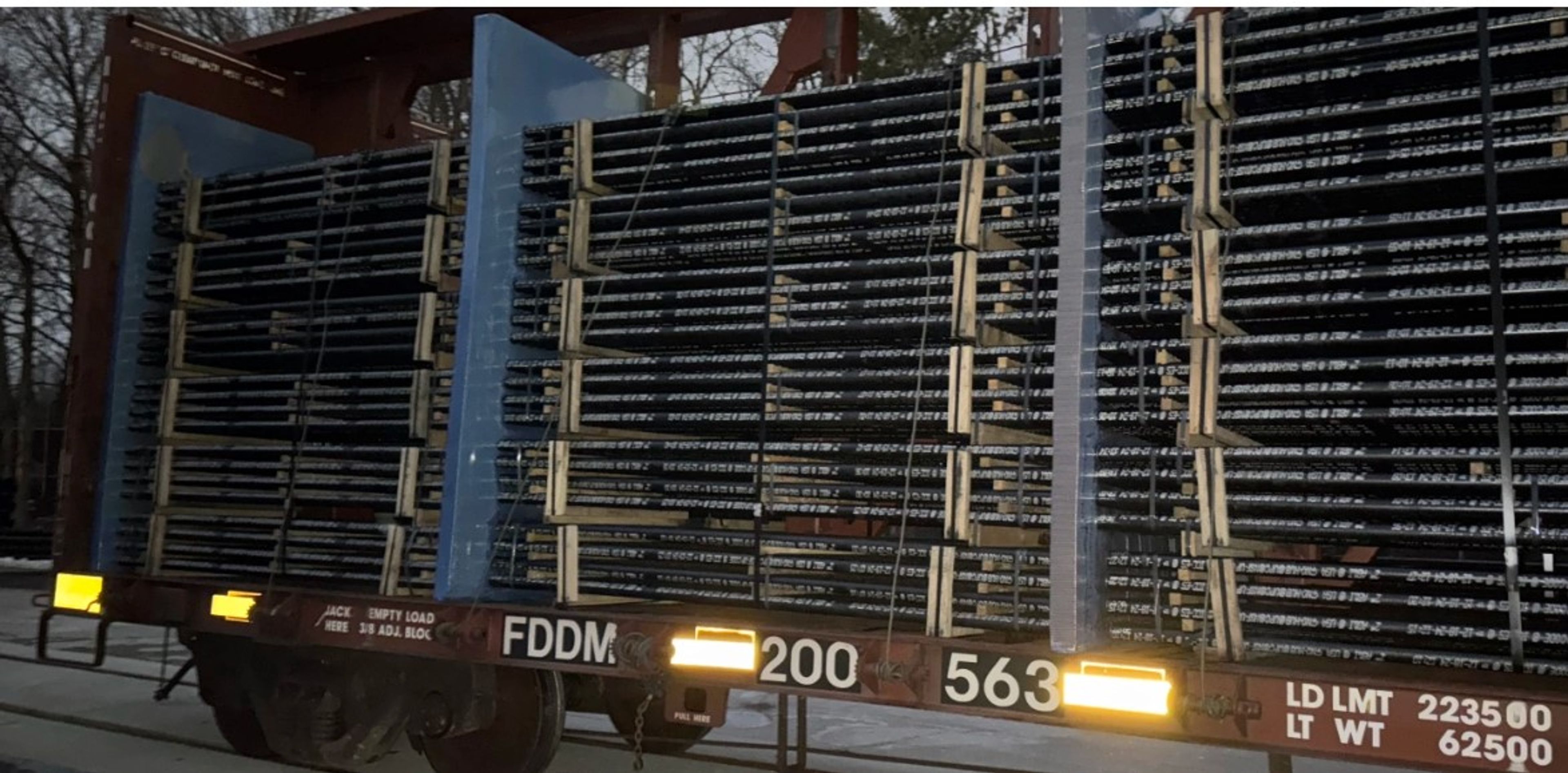 A railcar loaded with neatly stacked bundles of long metal rods secured upright, seen outdoors in low light. The railcar has visible markings, including “FDDM 200563” and weight limits.