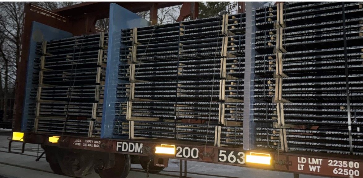 A railcar loaded with neatly stacked bundles of long metal rods secured upright, seen outdoors in low light. The railcar has visible markings, including “FDDM 200563” and weight limits.