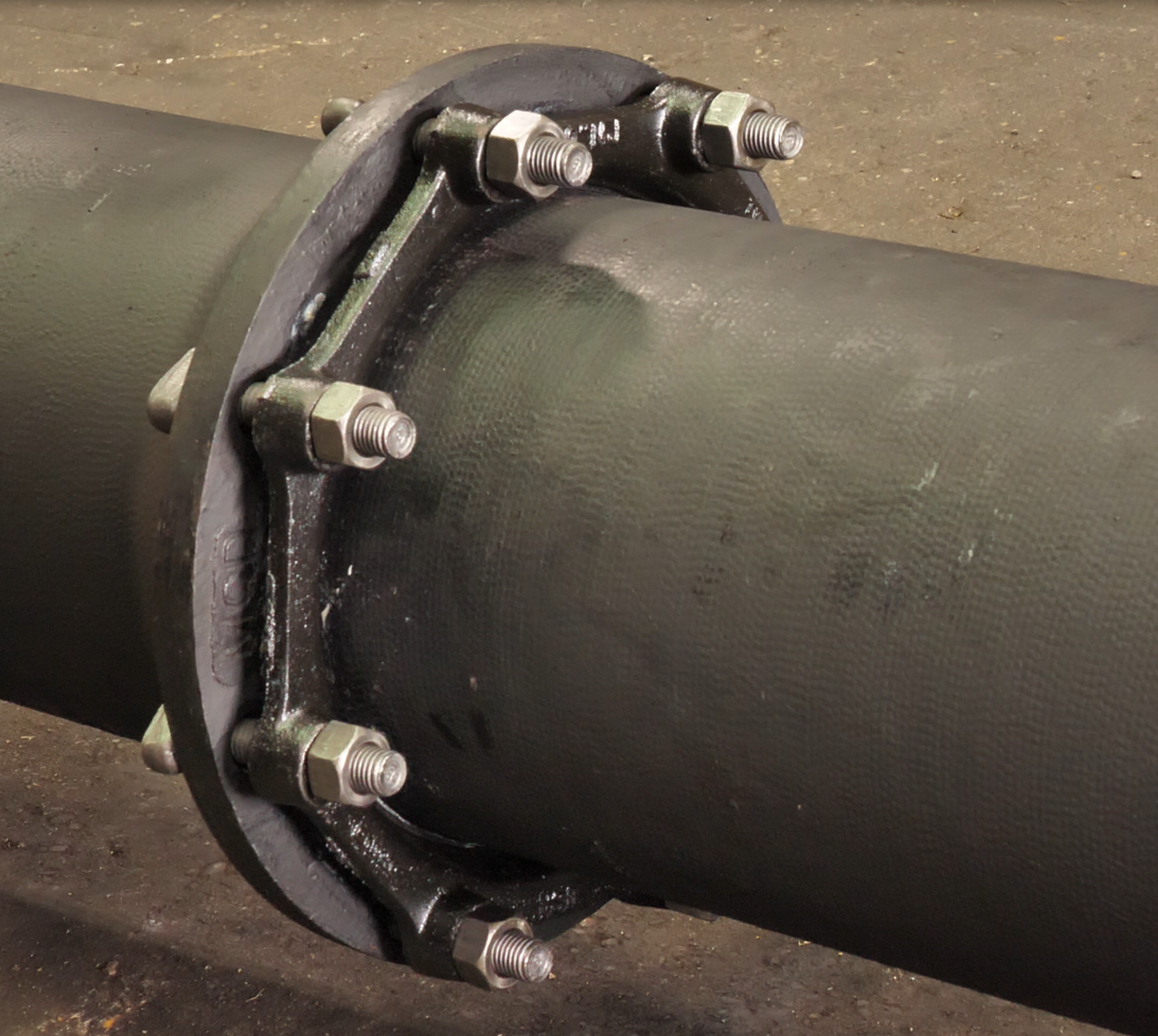A close-up view of a dark metal pipe with a flange joint secured by six large bolts and nuts, set against a rough, industrial floor background.