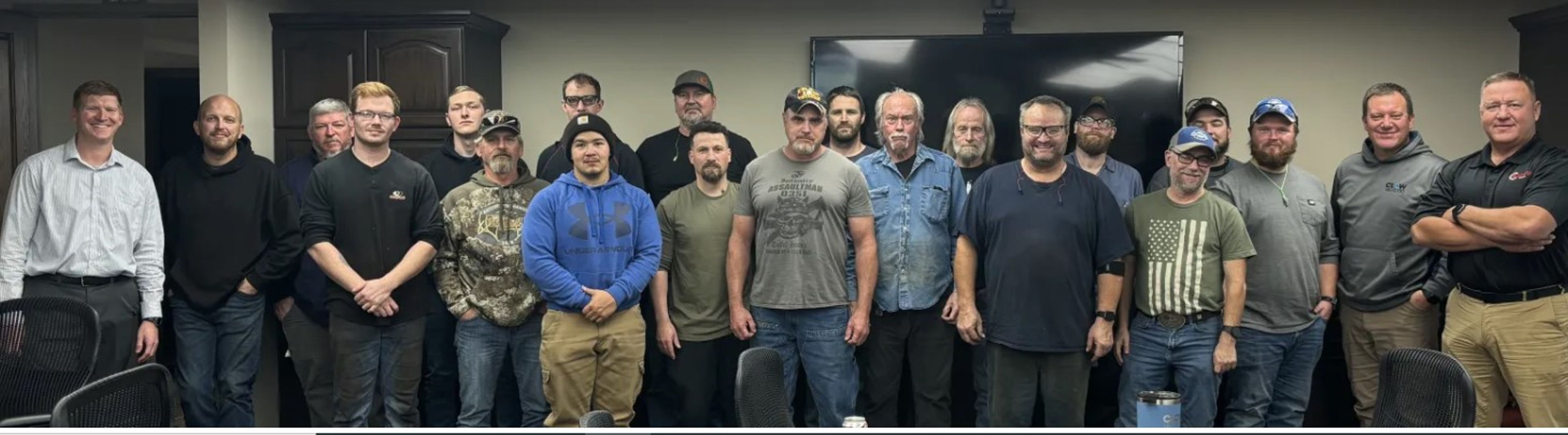 A group of twenty men stand together indoors, posing for a photo in a meeting room, with chairs and a large TV screen visible in the background.