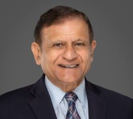 Smiling older man with short dark hair wearing a suit jacket, patterned tie, and light blue checked shirt, posed in front of a plain gray background.