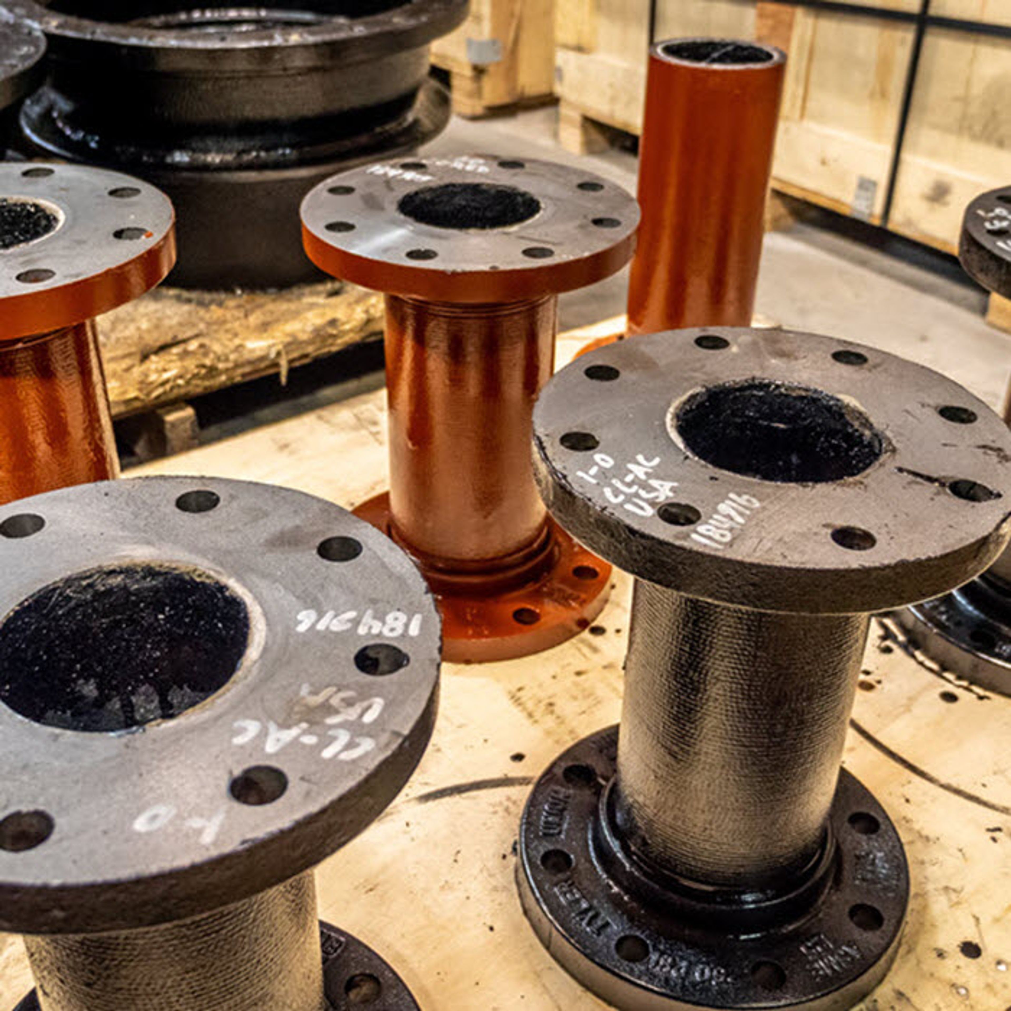 Several metal pipe flanges with handwritten markings are arranged upright on a wooden surface in an industrial setting, with more pipes and piping materials in the background.