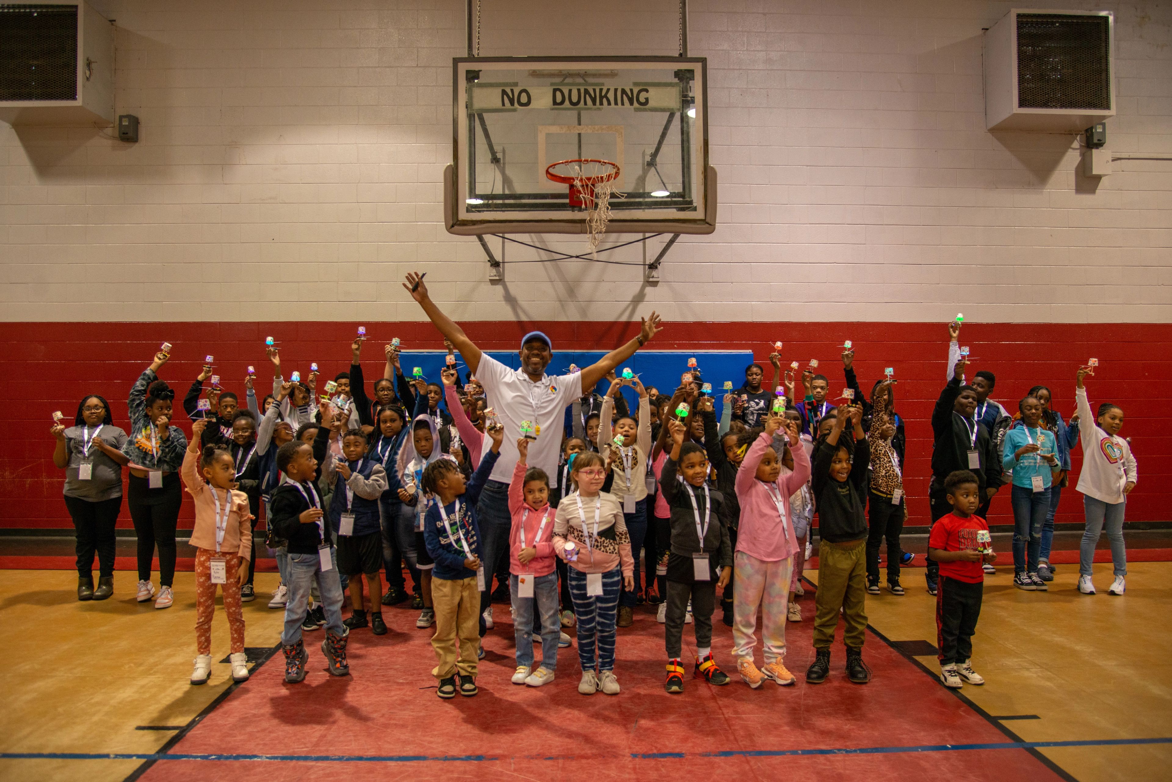 A group of children and adults pose in a gym under a basketball hoop, many raising small lights, with one person standing front and center with arms raised.