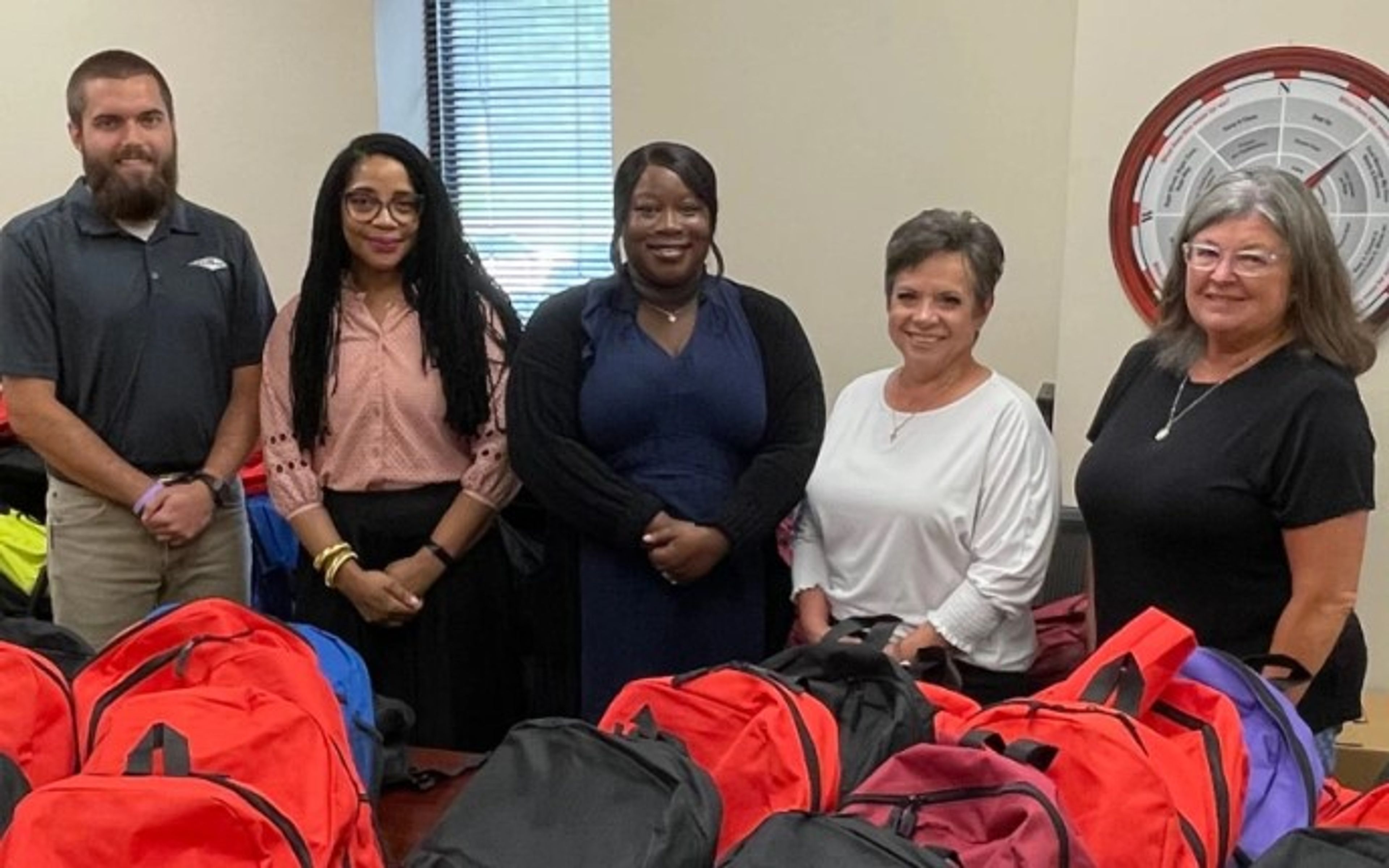 Five adults stand behind a table covered with colorful backpacks in a room with beige walls and a window. They are smiling and appear to be participating in a community or school supply event.