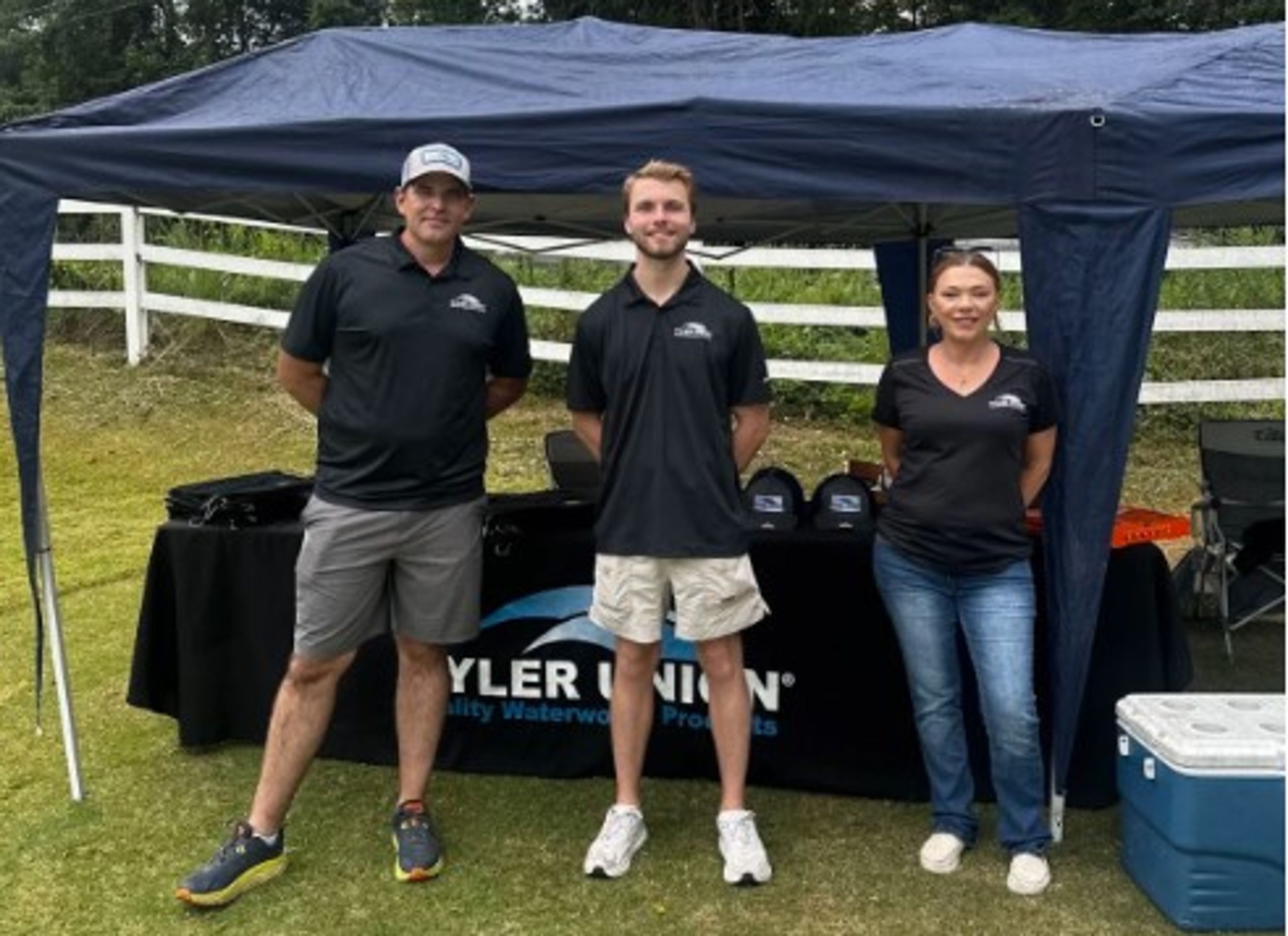 Three people stand under a canopy at an outdoor event with a “Tyler Union” table display behind them. They wear black shirts and look at the camera, with a cooler and white fence in the background.