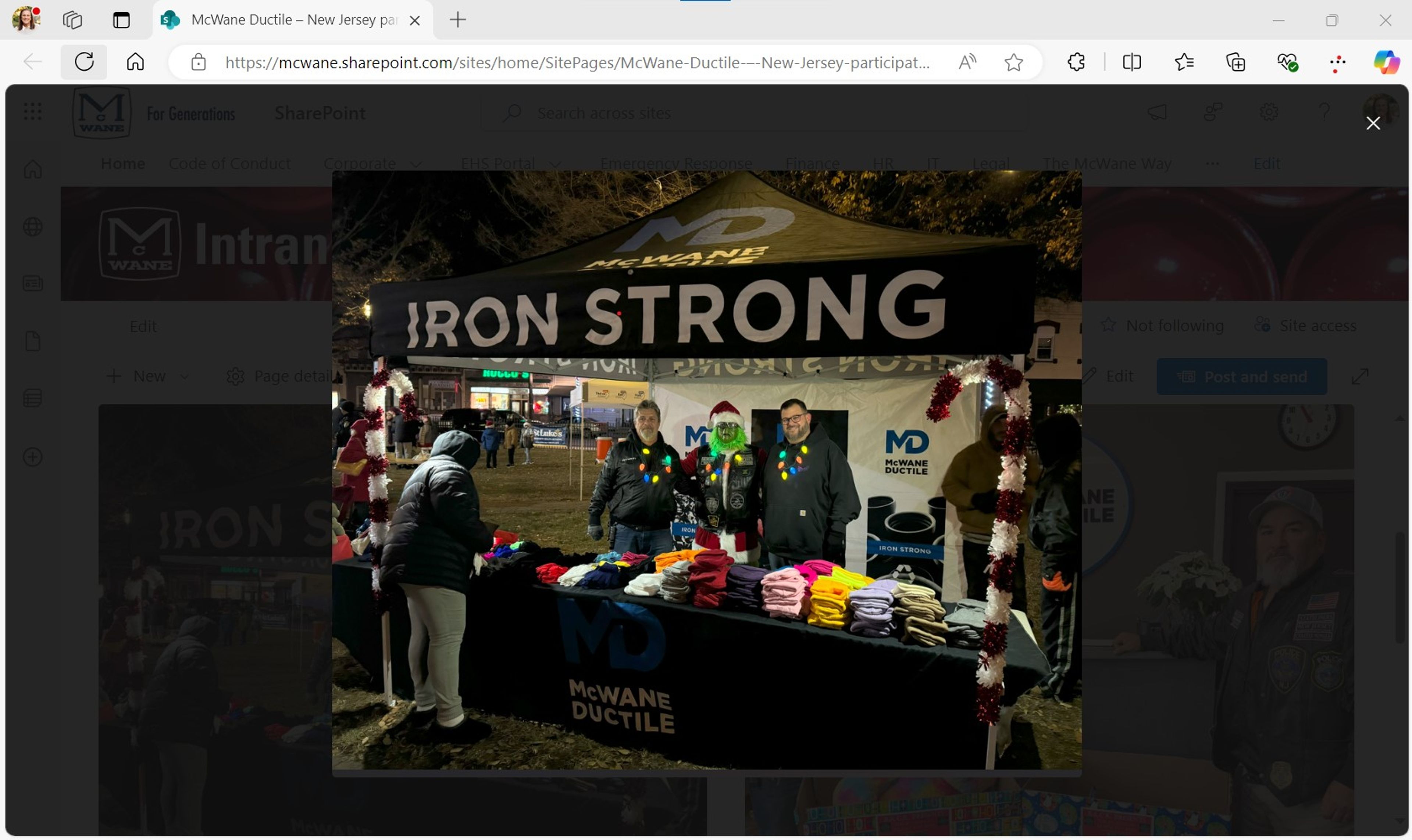 Four people stand behind a “IRON STRONG” McWane Ductile tent at an outdoor event, handing out colorful hats and scarves on a table, while several people collect items in front.