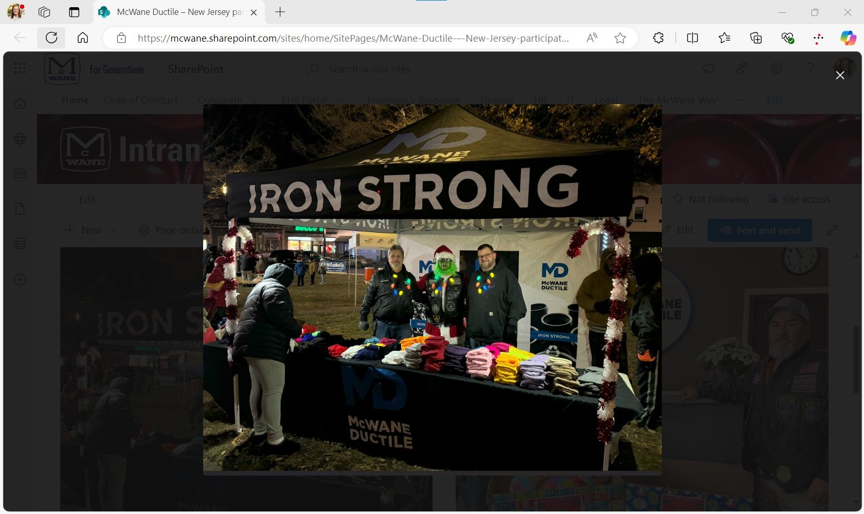 Four people stand behind a “IRON STRONG” McWane Ductile tent at an outdoor event, handing out colorful hats and scarves on a table, while several people collect items in front.