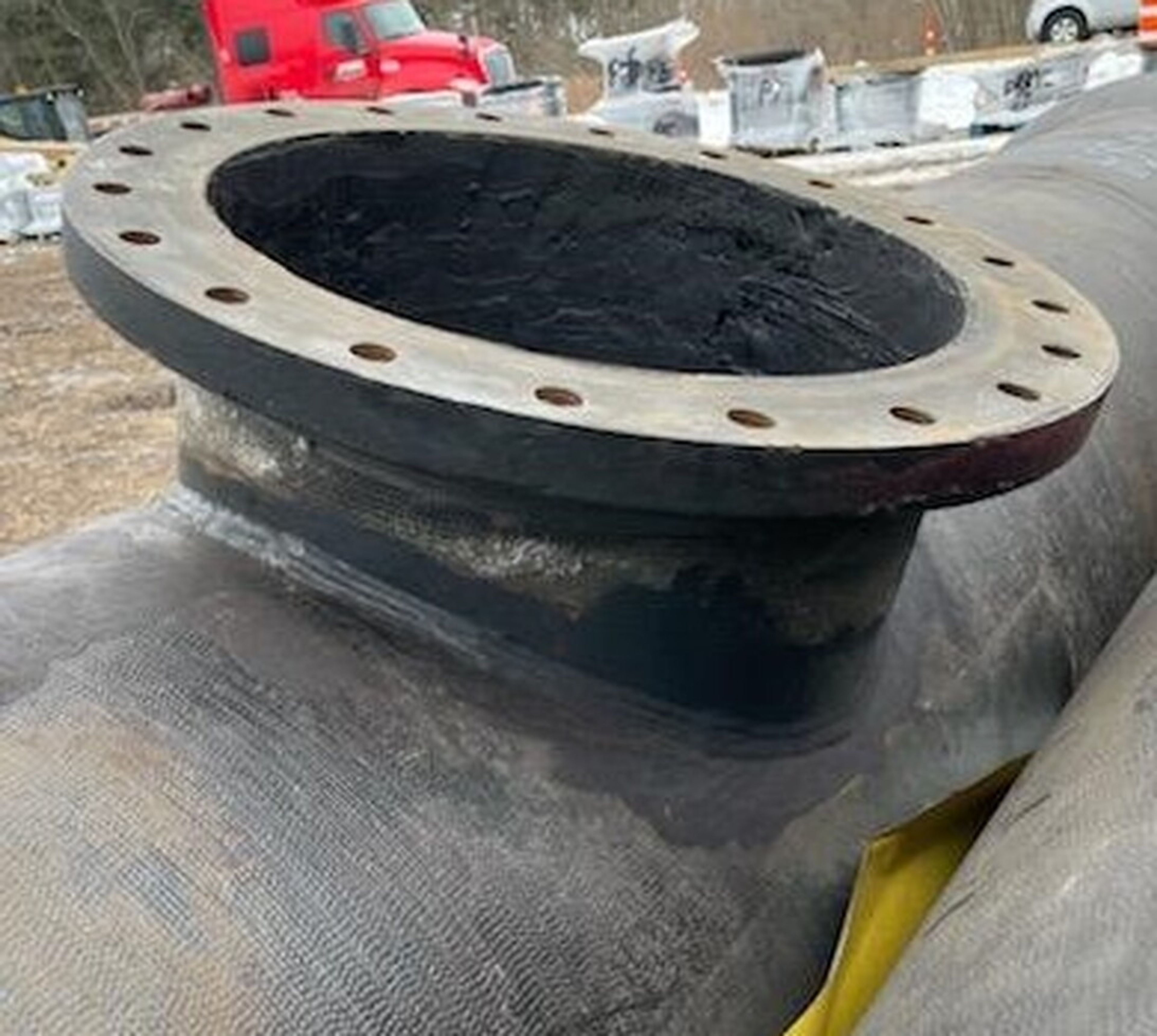 Large black metal pipe with a flanged circular opening, lying outdoors on the ground. The flange has several bolt holes around its edge. A red truck and construction materials are visible in the background.