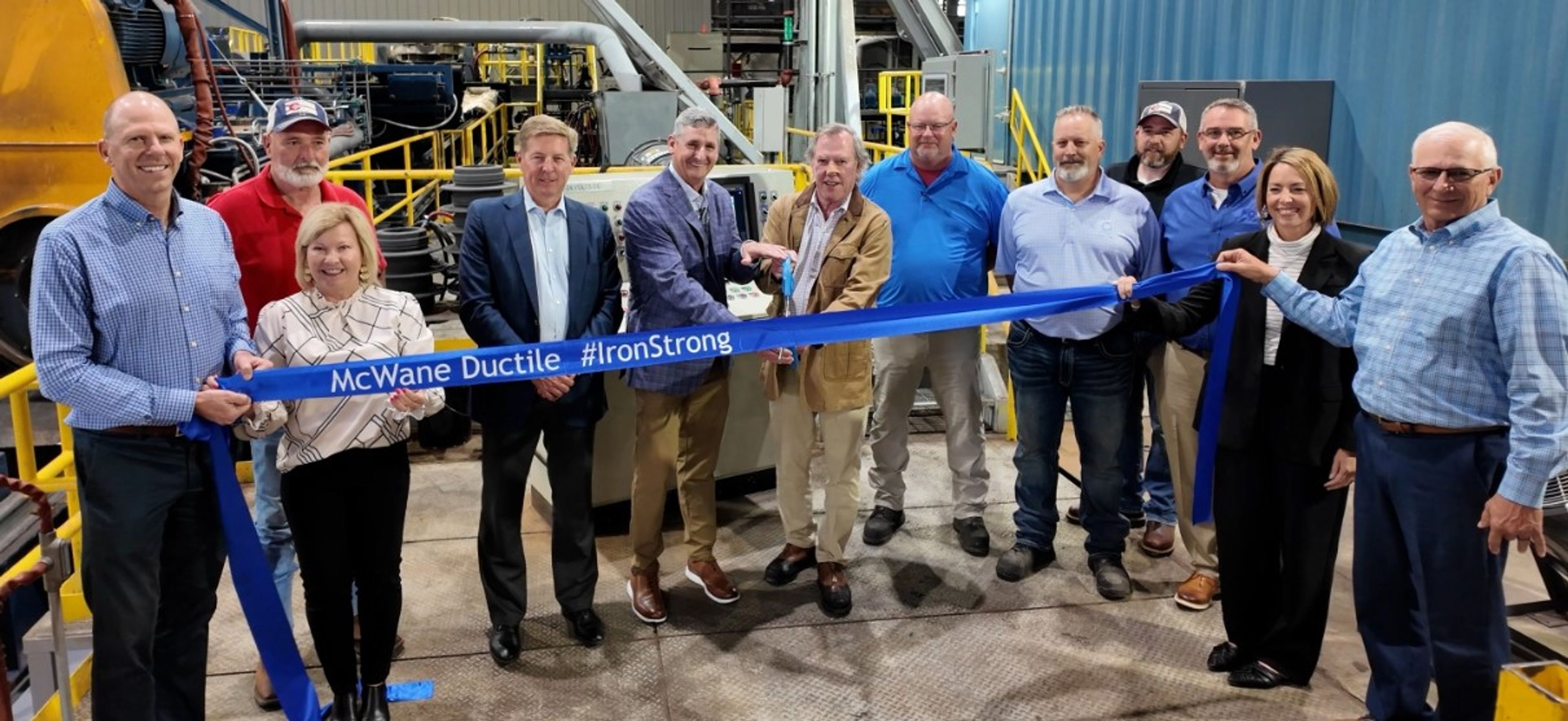 A group of people stand indoors holding a long blue ribbon that reads “McWane Ductile - #IronStrong” during a ribbon-cutting ceremony at an industrial facility.