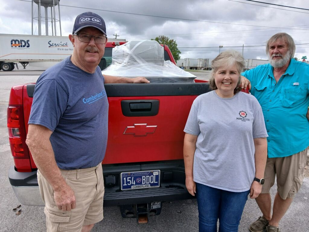 Three people stand behind a red Chevrolet pickup truck in a parking lot. The woman is smiling, and the two men stand on either side. A wrapped item is in the truck bed, and a water tower is visible in the background.