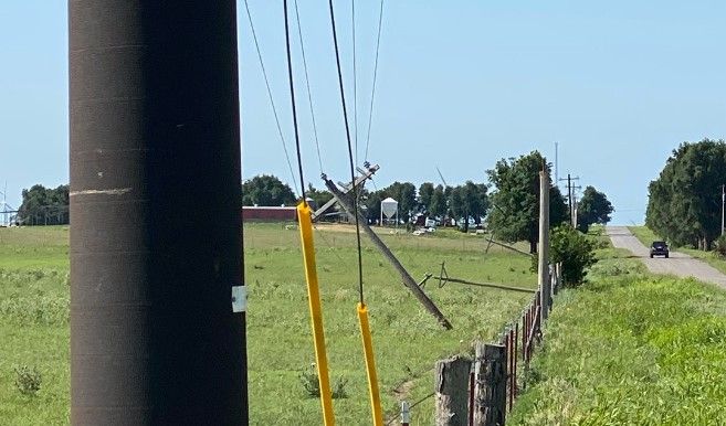 A rural scene with leaning power poles along a grassy roadside, one pole severely damaged and tilted, while a car drives down a distant roadway under a clear blue sky.