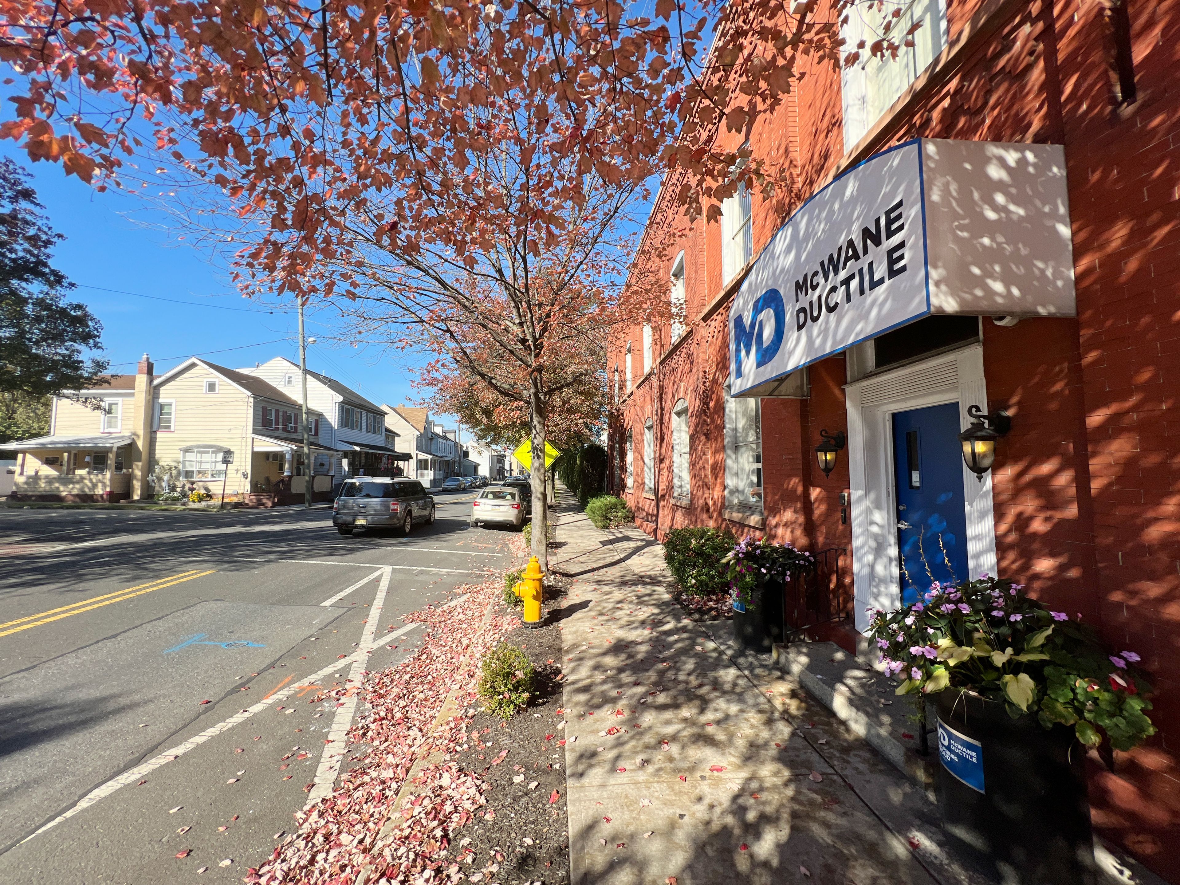 A sunny street scene shows autumn leaves on the sidewalk, a red brick building with a “McWane Ductile” sign, potted flowers, parked cars, and houses lining the street under a blue sky.