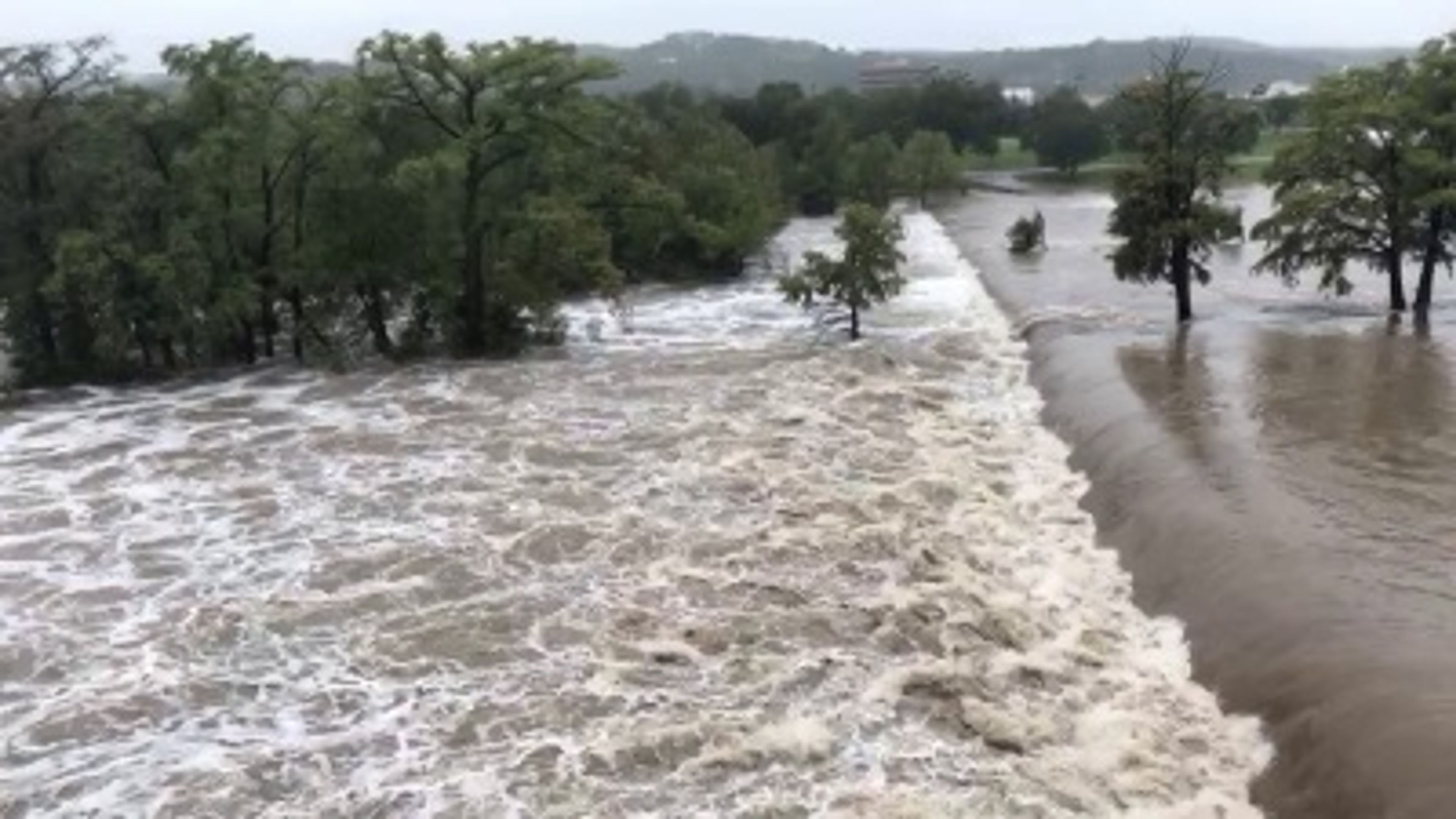 Floodwaters rush over a riverbank, submerging trees and surrounding land. The water is turbulent, and the area is visibly flooded, with only treetops above the waterline.