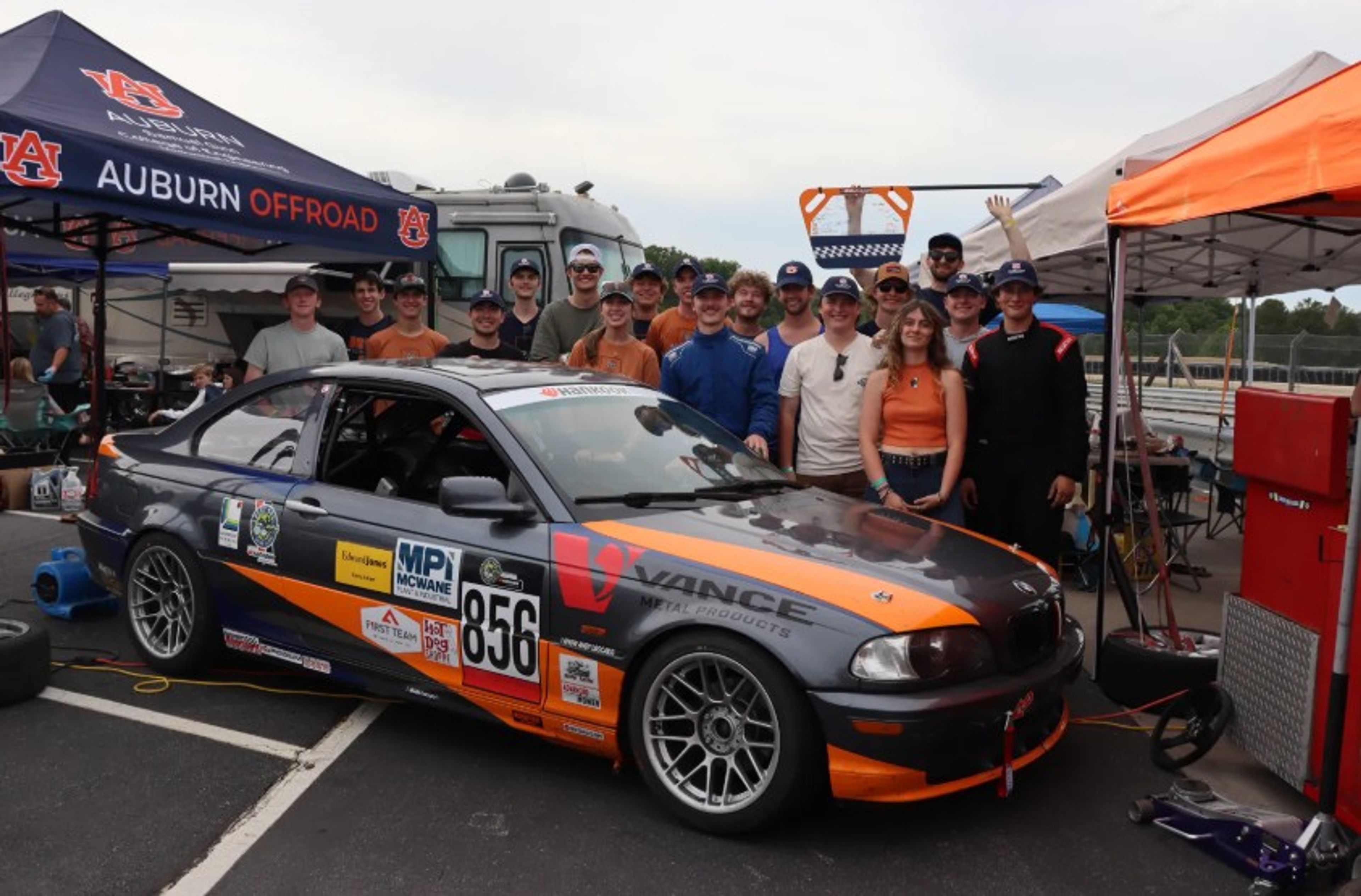 A group of people pose smiling behind a race car under tents labeled “Auburn Offroad” at a racetrack, with equipment and a trailer visible in the background.