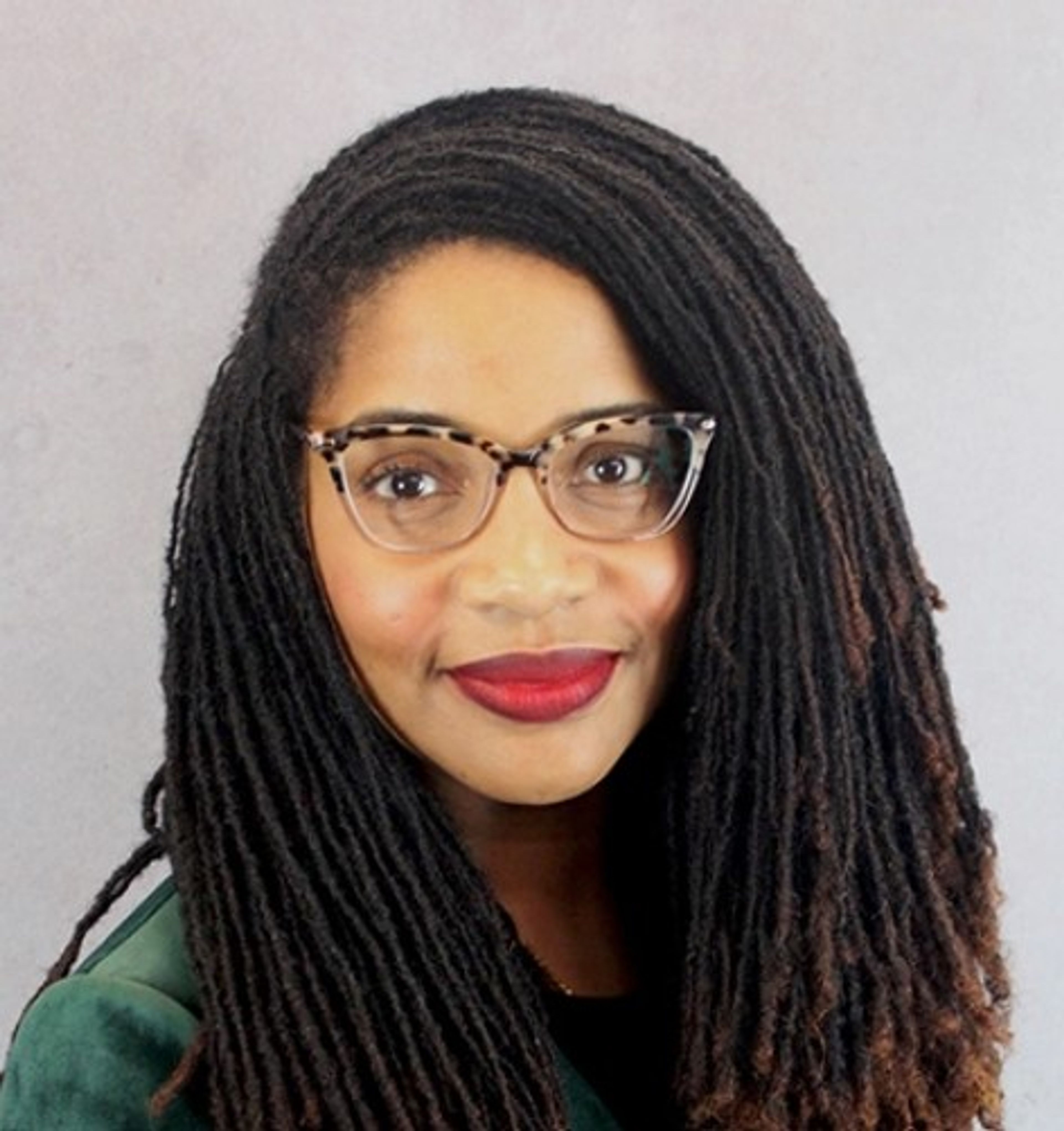 A woman with long, dark, twisted hair wears eyeglasses with leopard-print frames and red lipstick. She is smiling slightly and posing in front of a plain light gray background.