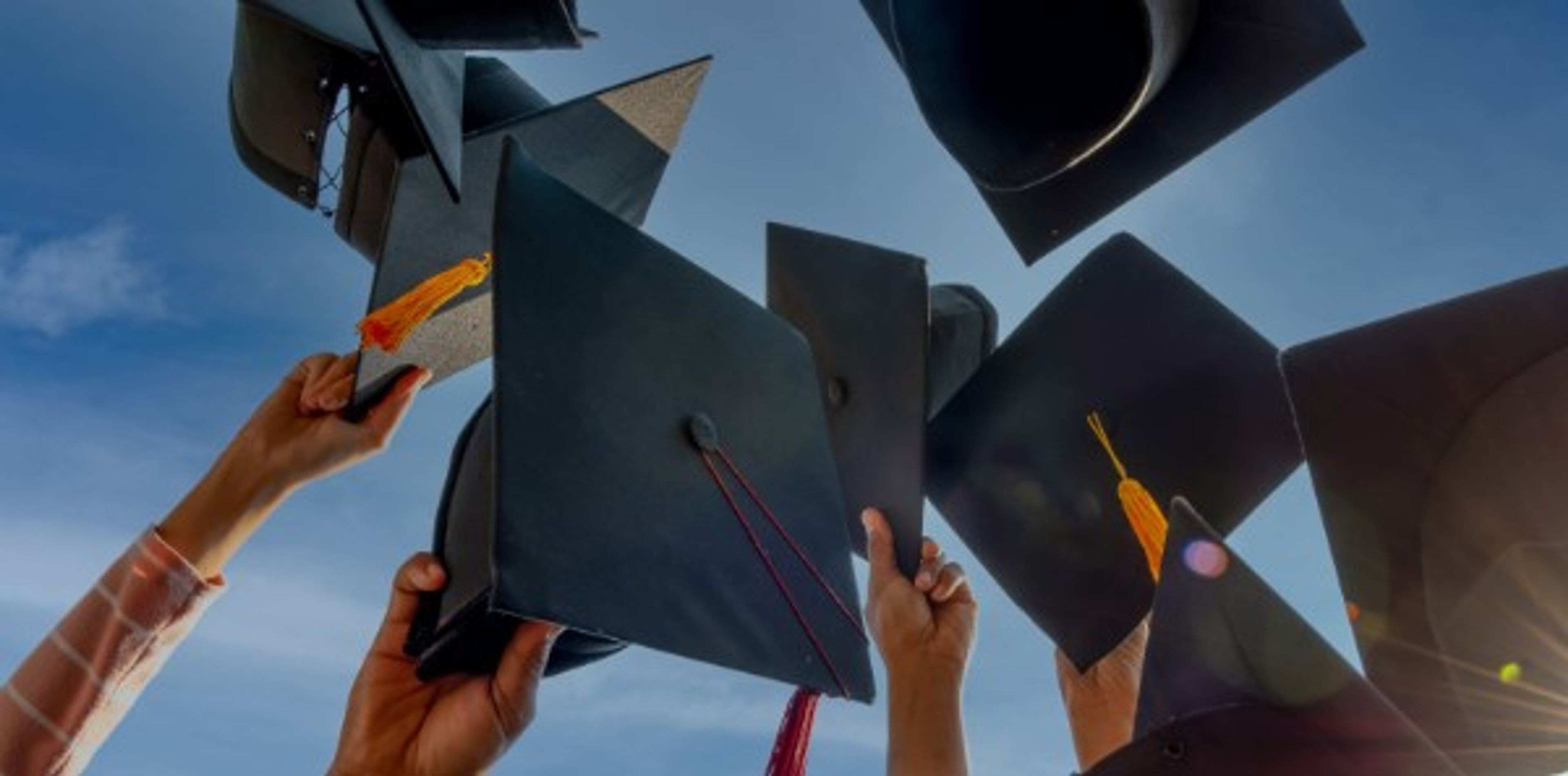 Several hands hold graduation caps up toward a blue sky, celebrating, with sunlight shining in the background.