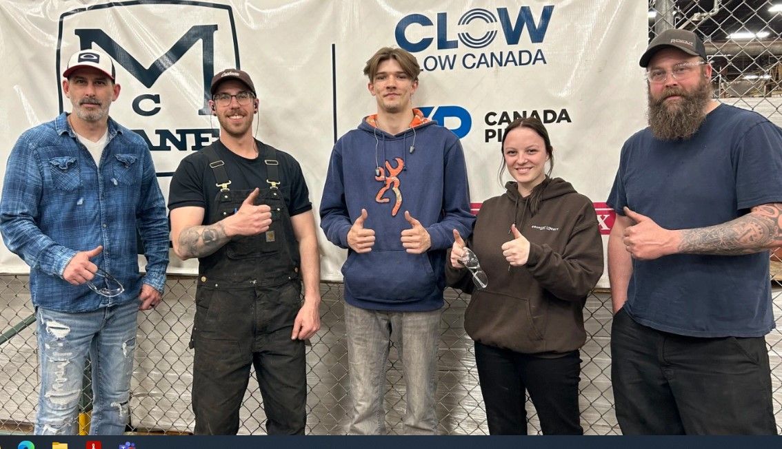 Five people stand in front of a banner with logos, all smiling and giving thumbs up. They wear casual clothing, including jeans, hoodies, and caps. The background includes a metal fence and parts of workshop equipment.