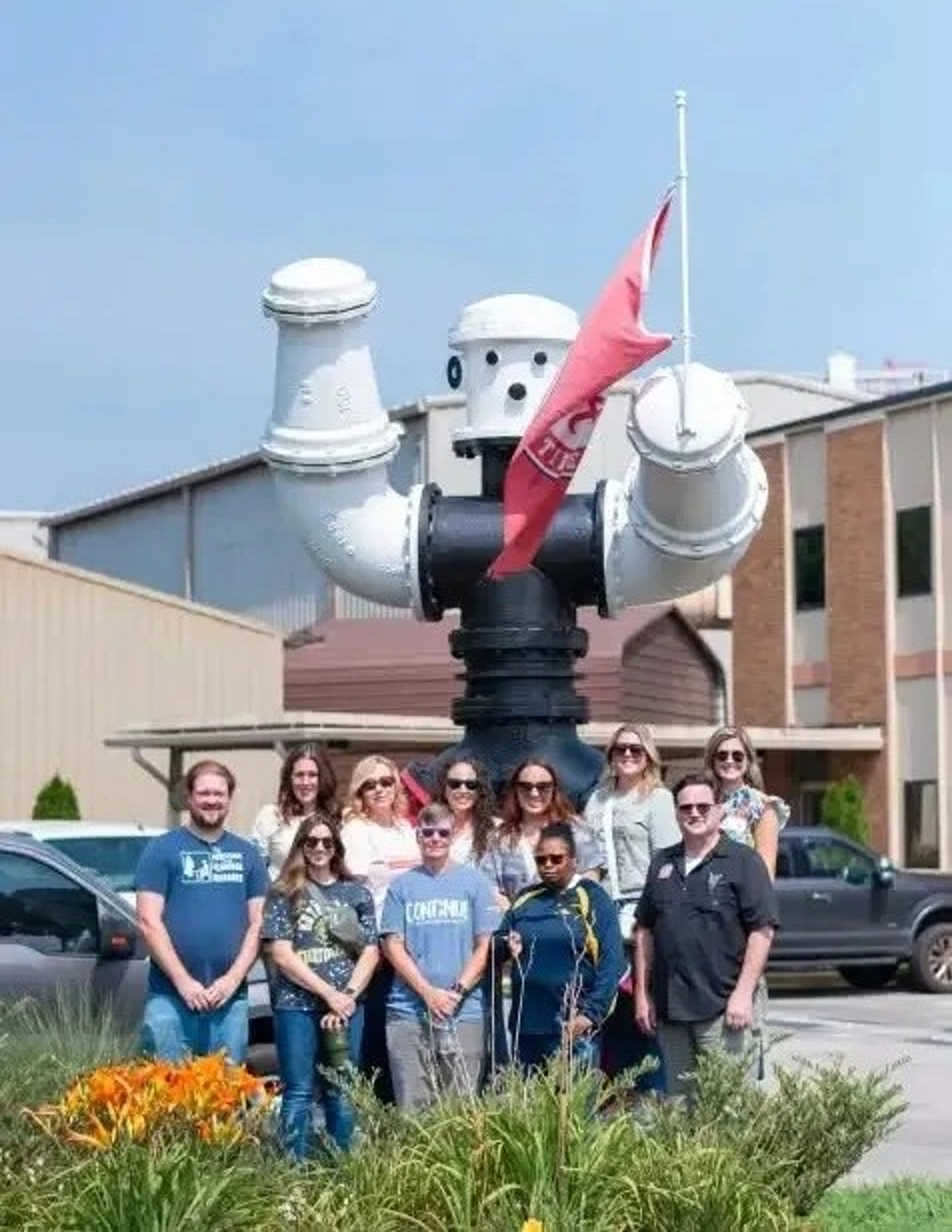 A group of people stands smiling in front of a large white robot statue holding a red flag, outside an industrial building on a sunny day.