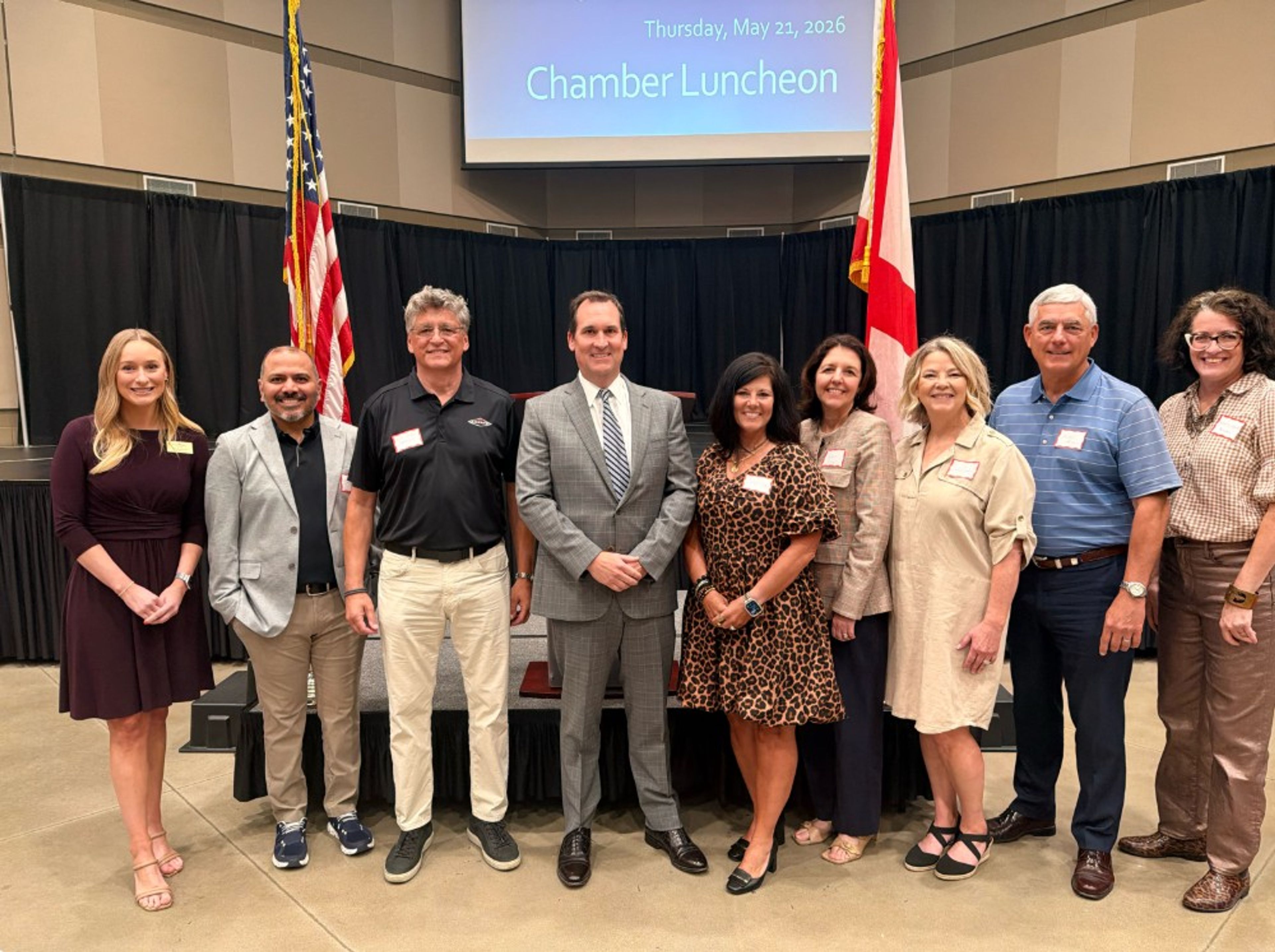 Nine people stand indoors in front of two flags and a screen reading “Chamber Luncheon.” They are dressed in business and business-casual attire, smiling at the camera.