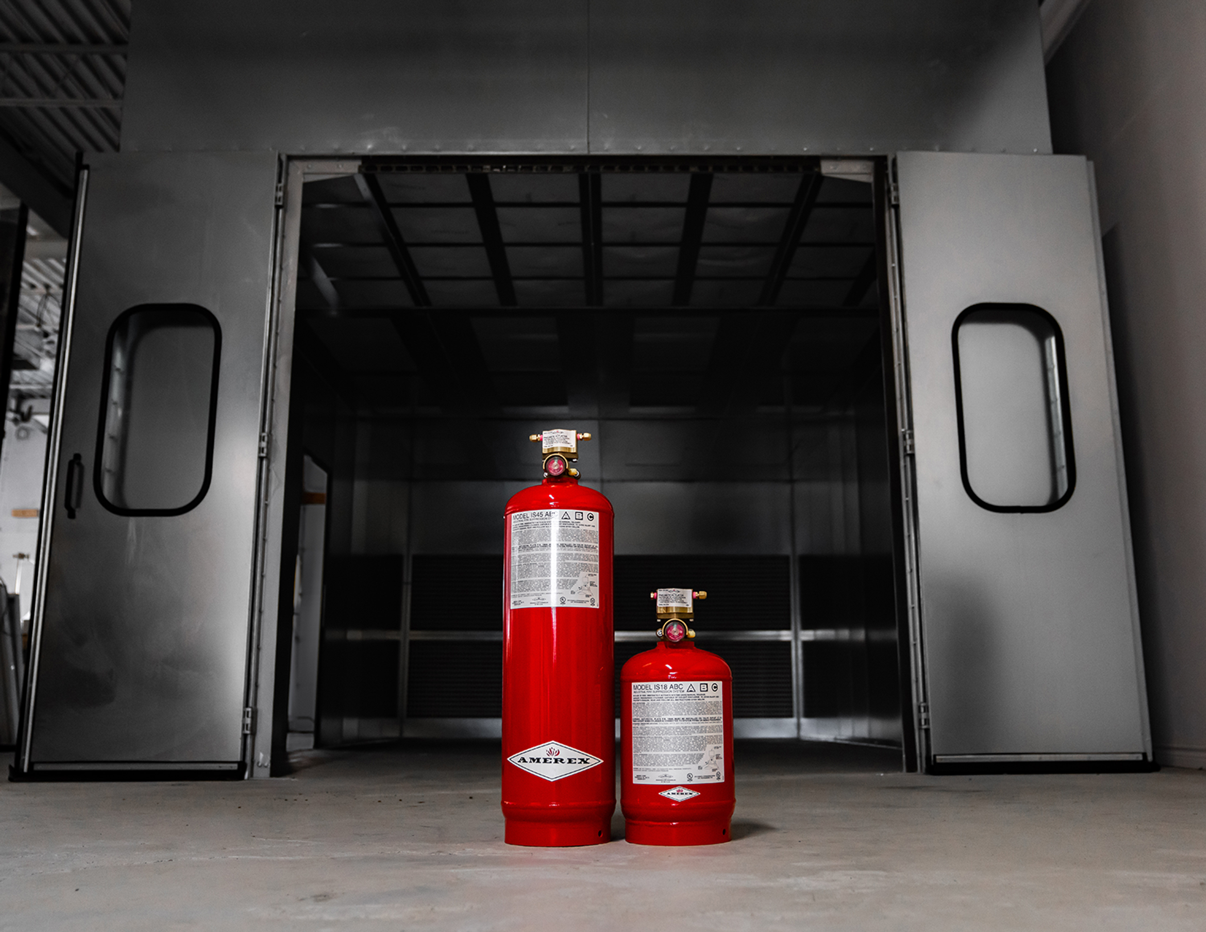 Two red fire extinguishers, one large and one small, stand on a gray floor in front of a large metallic industrial door with black handles.