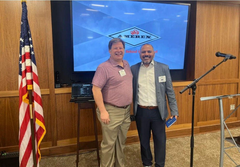 Two men smiling and standing in front of a large Amerex display screen in a wood-paneled room with an American flag and a microphone stand nearby.