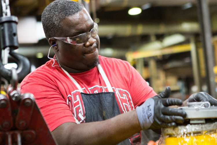 A man wearing safety glasses, gloves, and a red shirt works with machinery in an industrial setting. The background is blurred with equipment visible.
